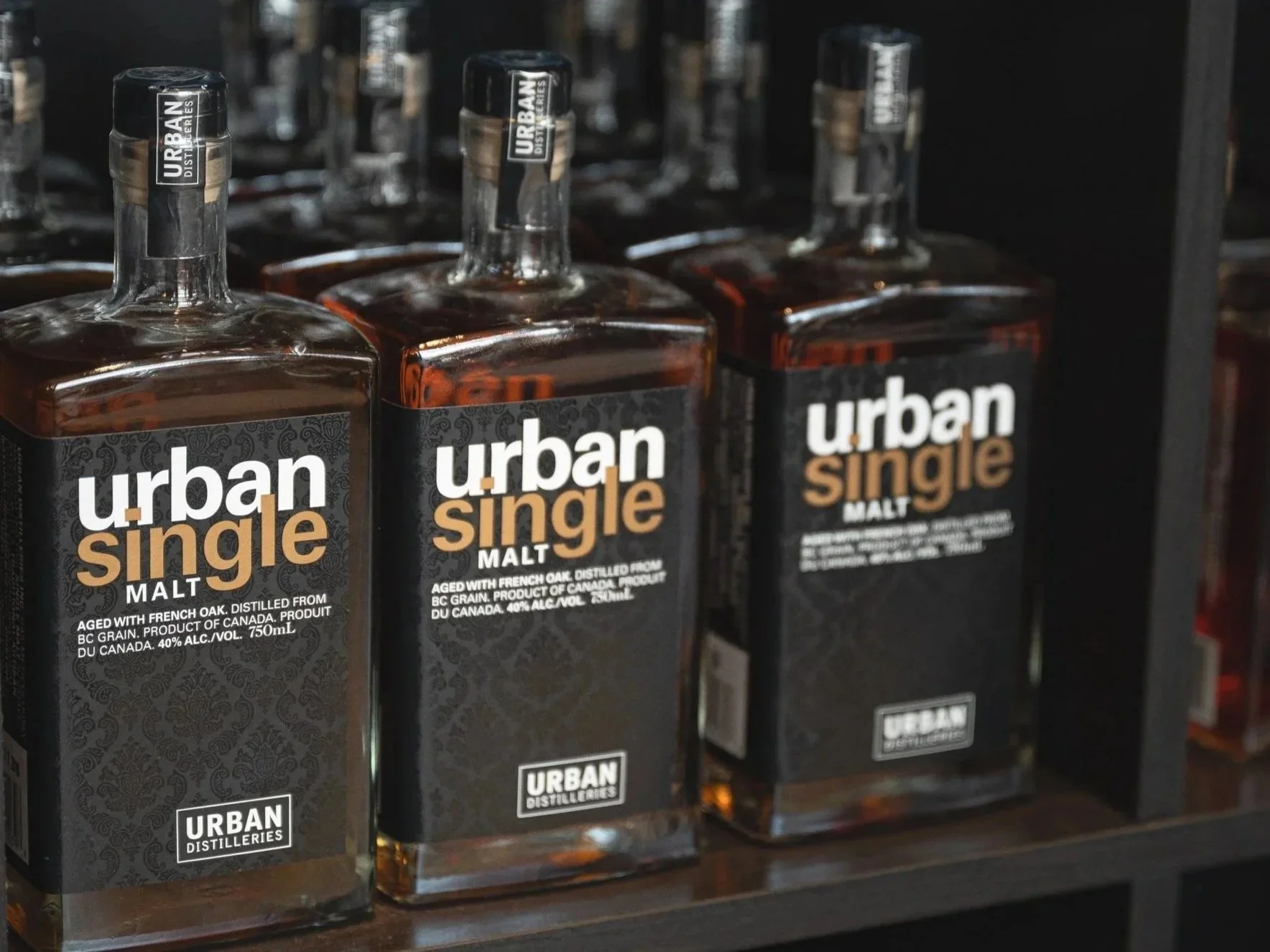 Multiple bottles of Urban Single Malt whiskey on a shelf. The labels indicate it is aged with French oak, distilled from BC grain, and produced in Canada with 40% alcohol content.