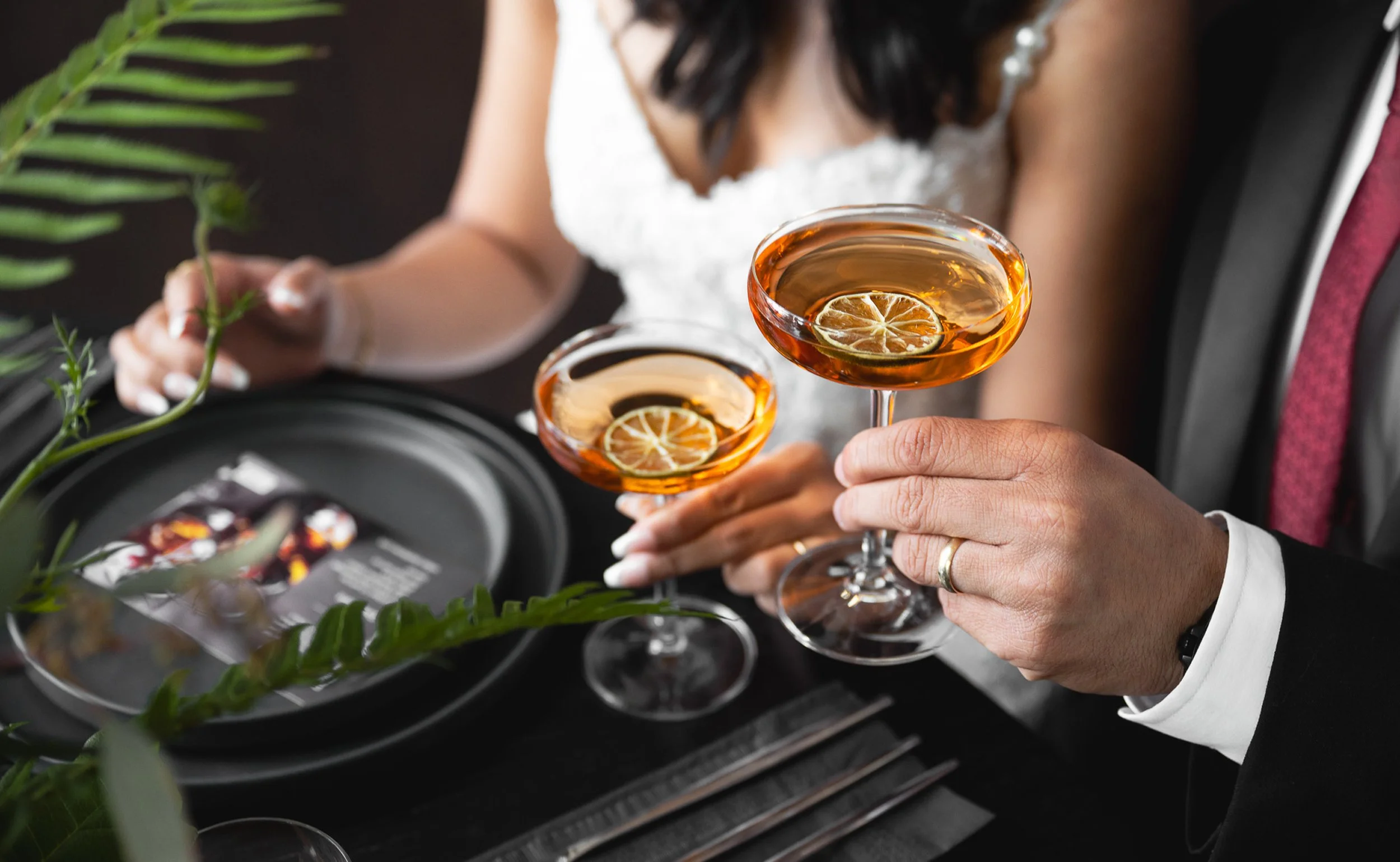Bride and groom holding handcrafted orange cocktails garnished with lemon slices at their wedding dinner table.