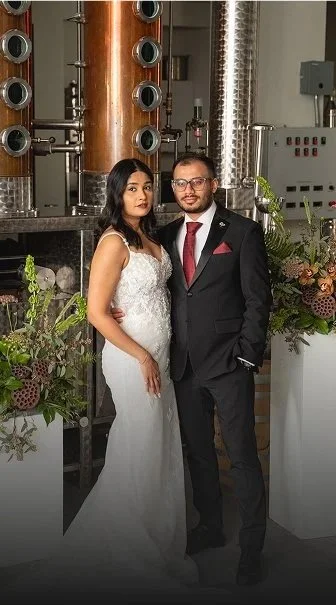 A newlywed couple stands together at a floral alter, surrounded by industrial machinery in a distillery