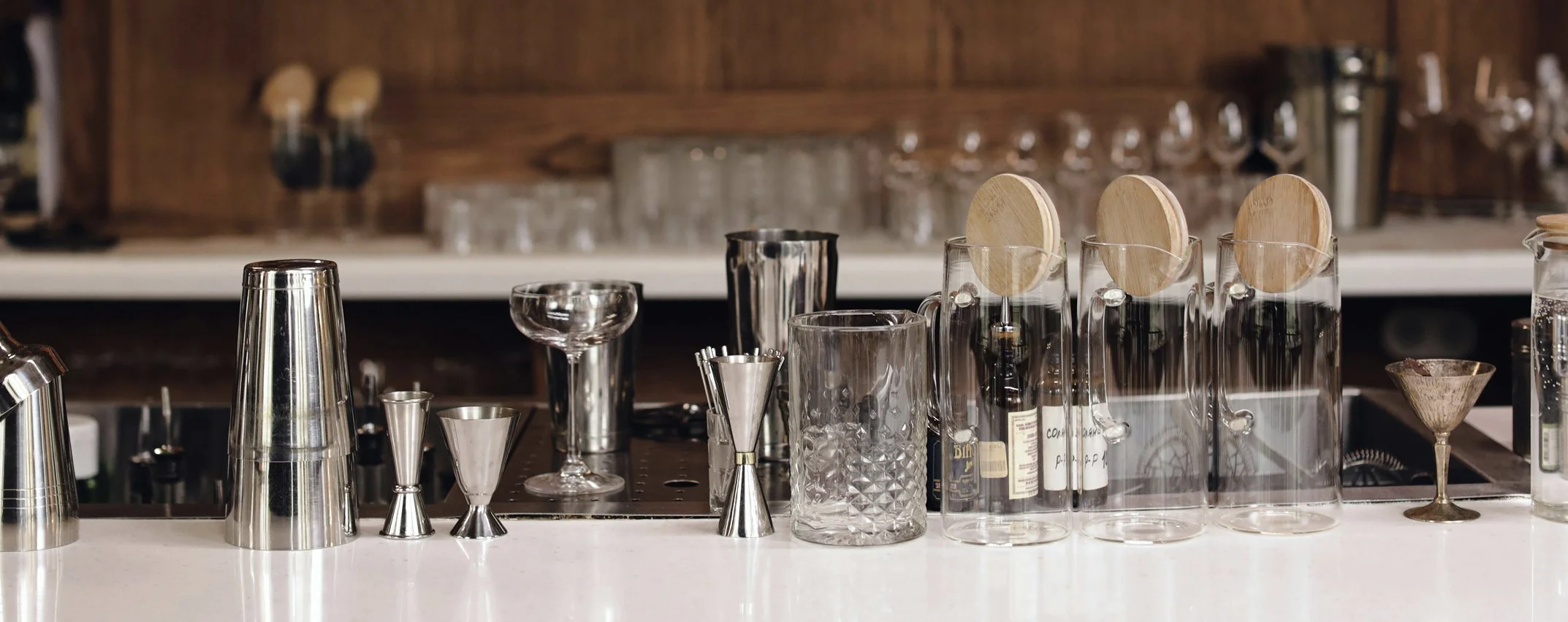 Bar counter with various barware including glasses, shakers, jiggers, and wooden stir sticks, with glassware on the shelves in the background.