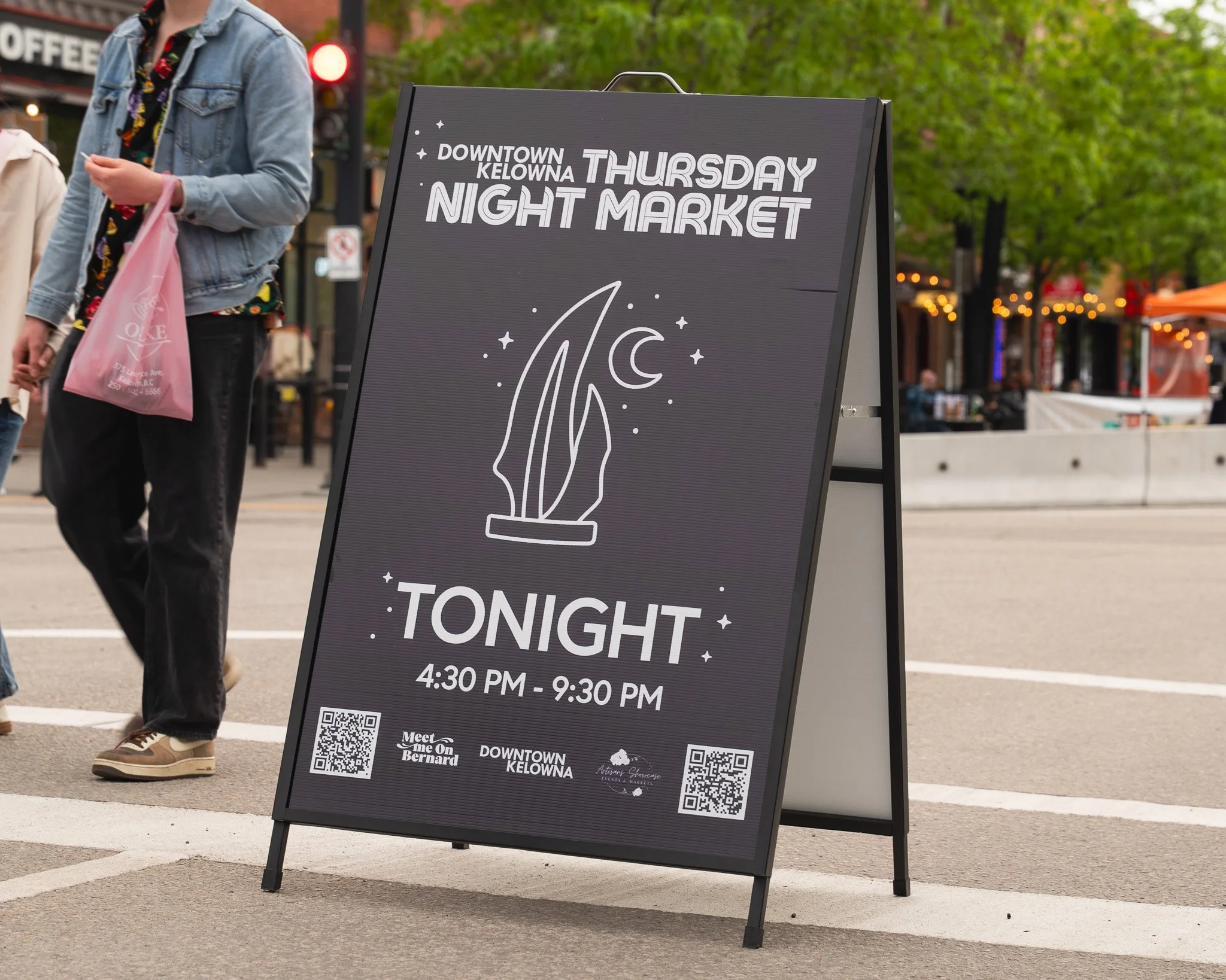A black sidewalk signboard advertising a downtown Kelowna Thursday night market from 4:30 PM to 9:30 PM, with a graphic of the Sails statue and QR codes at the bottom.