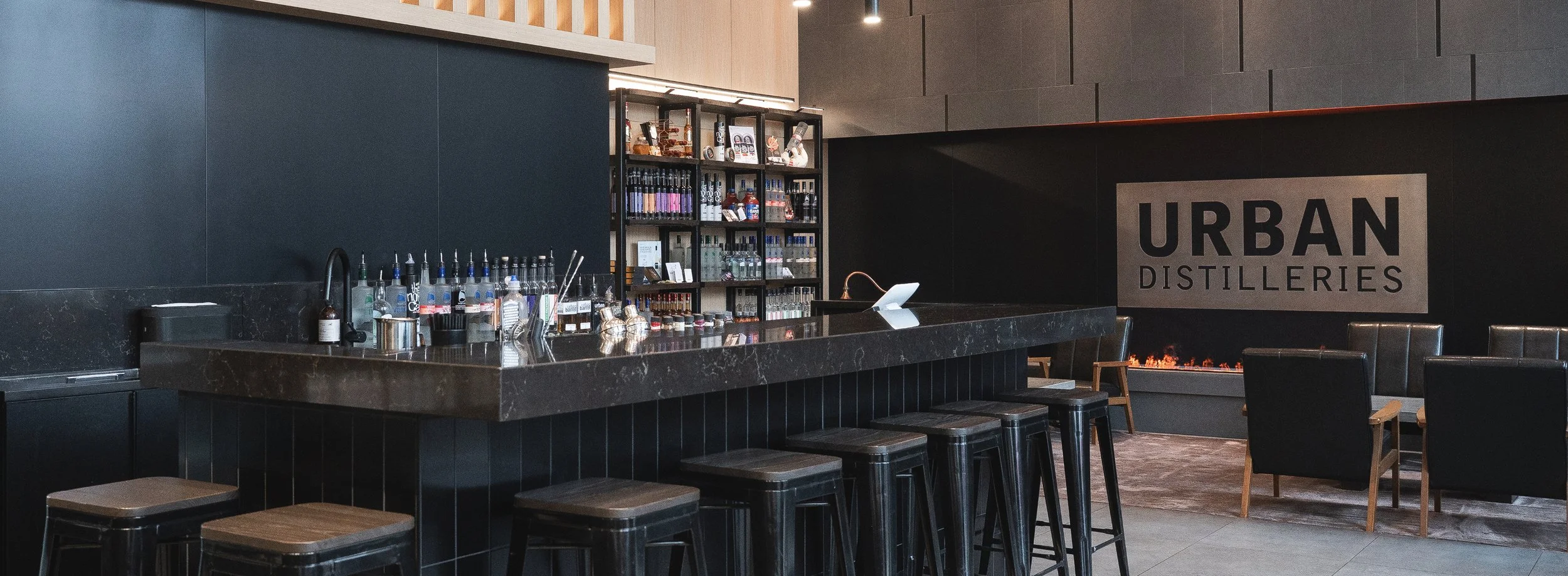 Modern bar with a black marble countertop, liquor display, and bar seating, overlooking a lounge featuring an Urban Distilleries sign and a contemporary fire feature.