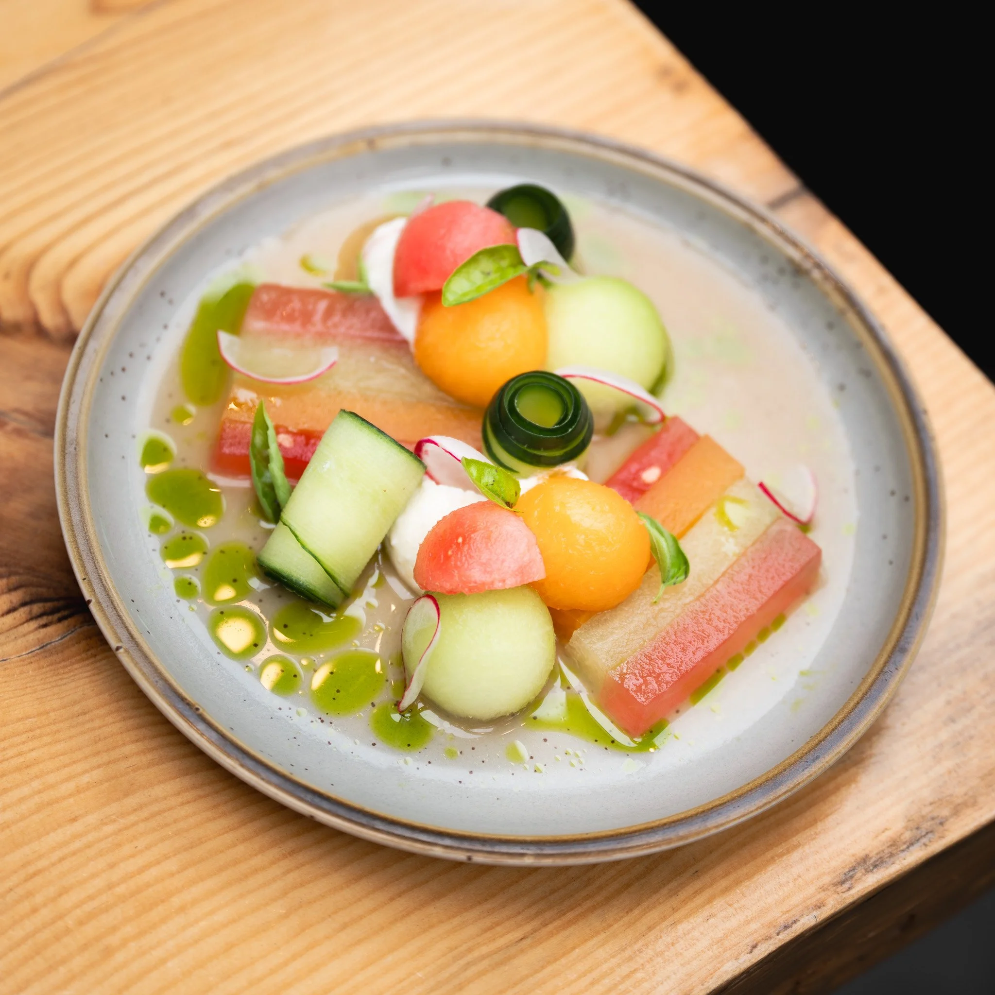 A melon salad with sliced radishes, rolled cucumber, melon balls, whipped feta, and herbs, drizzled with green oil, in a bowl on a wooden table.