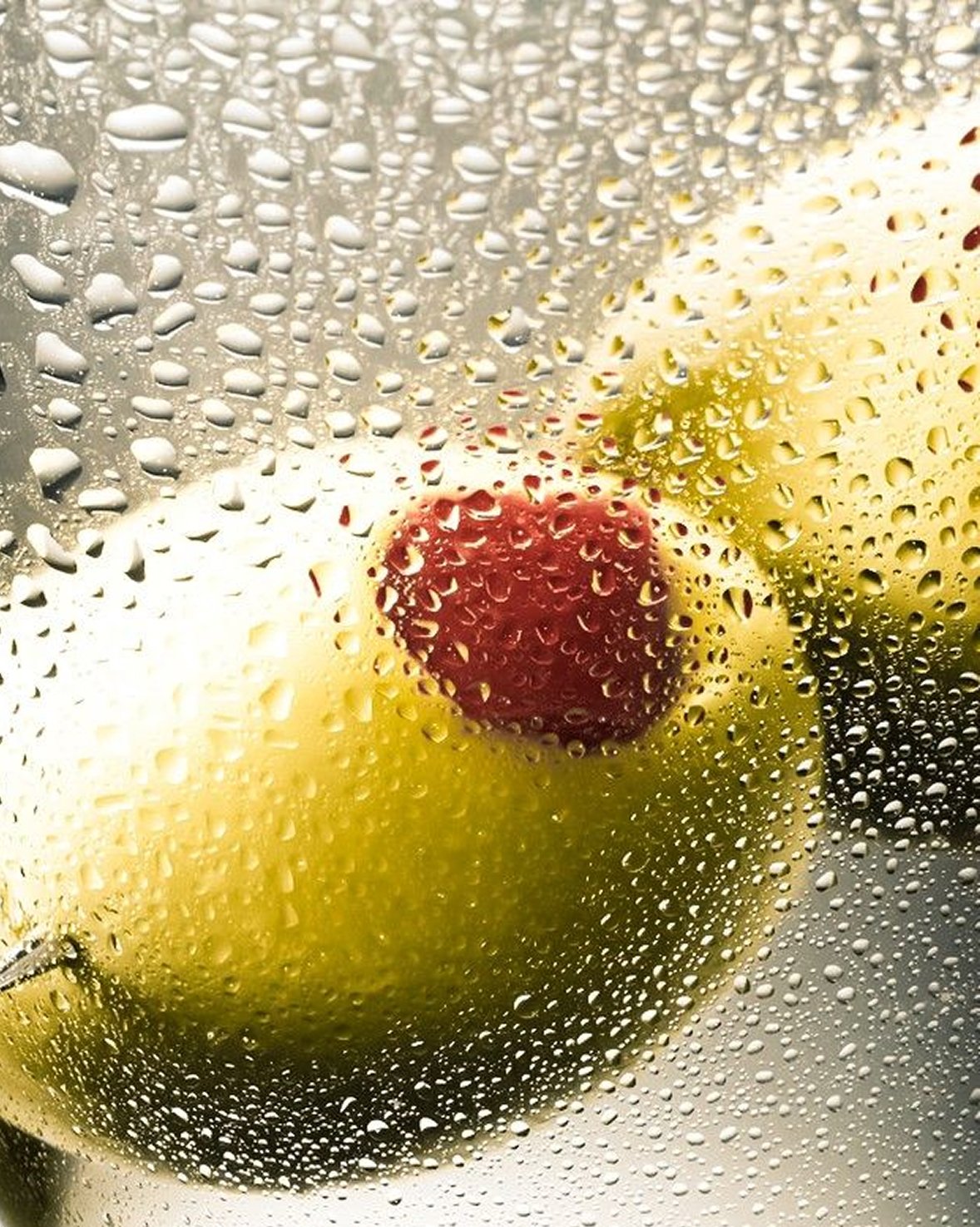 A green olive stuffed with a red pepper seen through a foggy martini glass covered with water droplets.