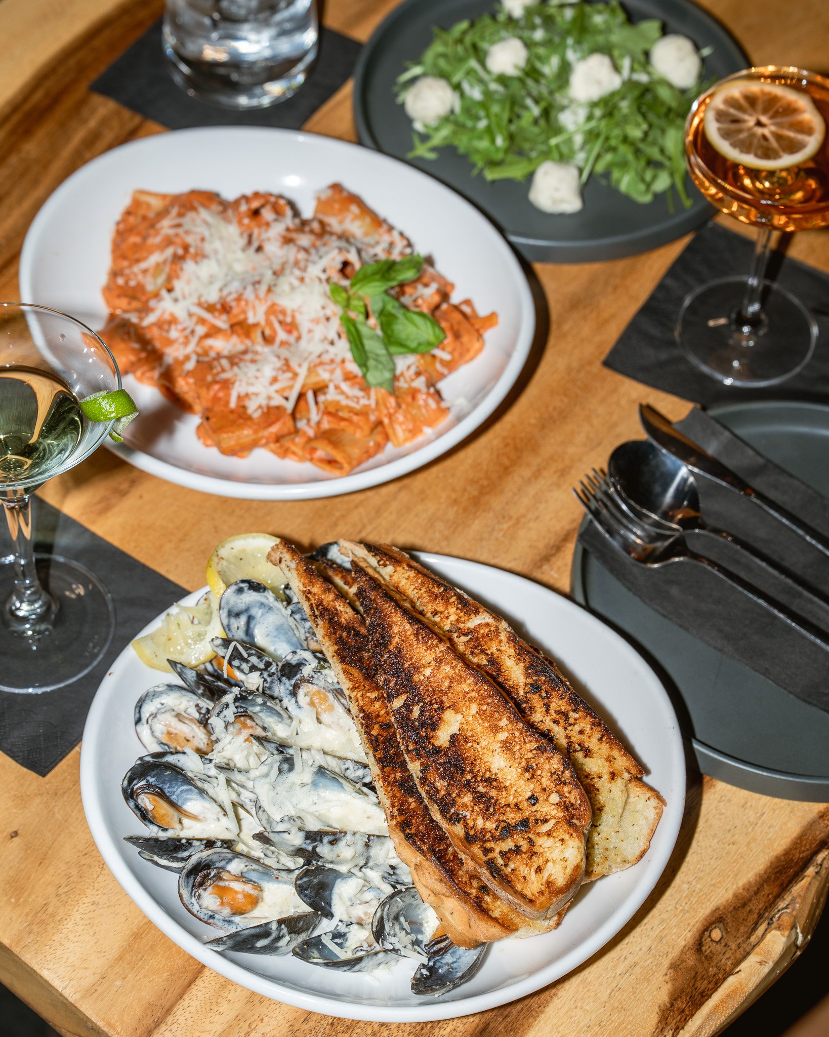 Restaurant meal featuring mussels in a creamy sauce with focaccia, pasta topped with tomato sauce and grated cheese, a fresh salad, and refreshing beverages on a wooden table.