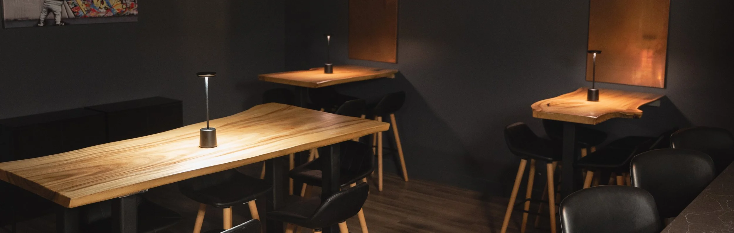 A dimly lit speakeasy interior with wooden tables, black chairs, and small modern table lamps, featuring dark walls and artwork.