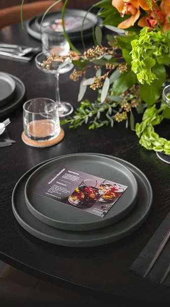 A dining table set with dark gray plates, a recipe card, and glasses, decorated with a large floral arrangement of various green leaves and flowers.