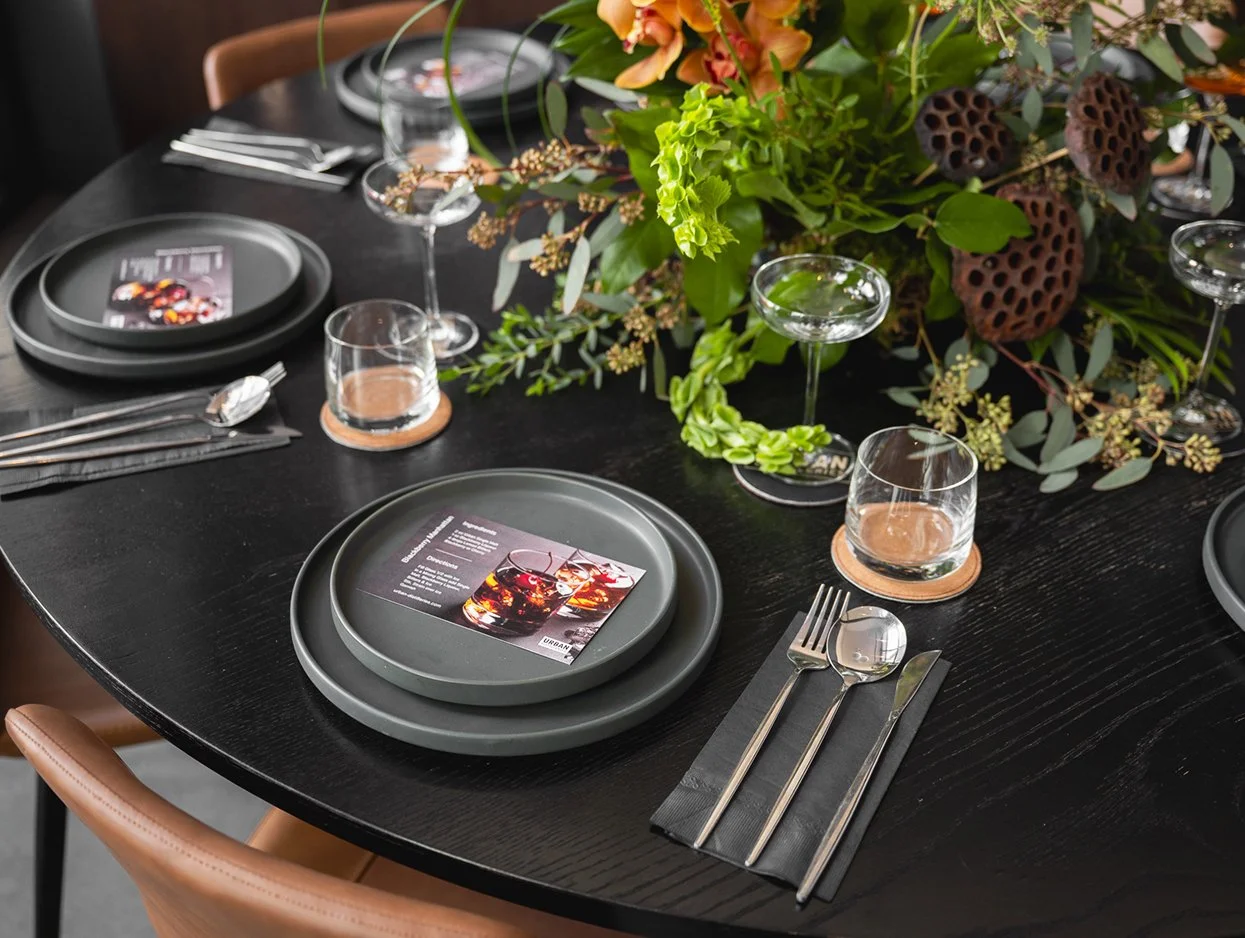 A black dining table set for a formal event with black plates, silverware, and drinking glasses. A large floral centerpiece with green leaves, orange orchids, and brown seed pods decorates the table.