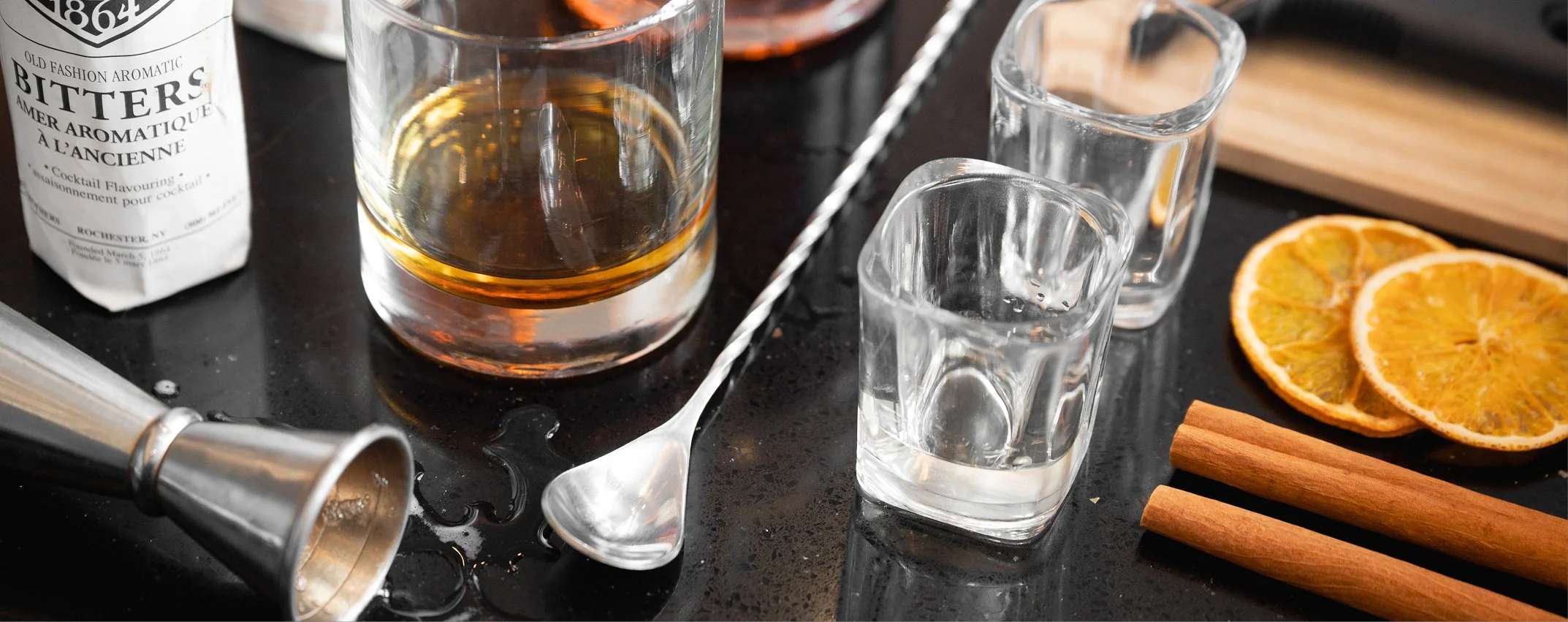 A bar countertop with a partially filled glass of whiskey, two shot glasses filled with water, dried orange slices, cinnamon sticks, a cocktail jigger, a bar spoon, and a bottle of bitters.