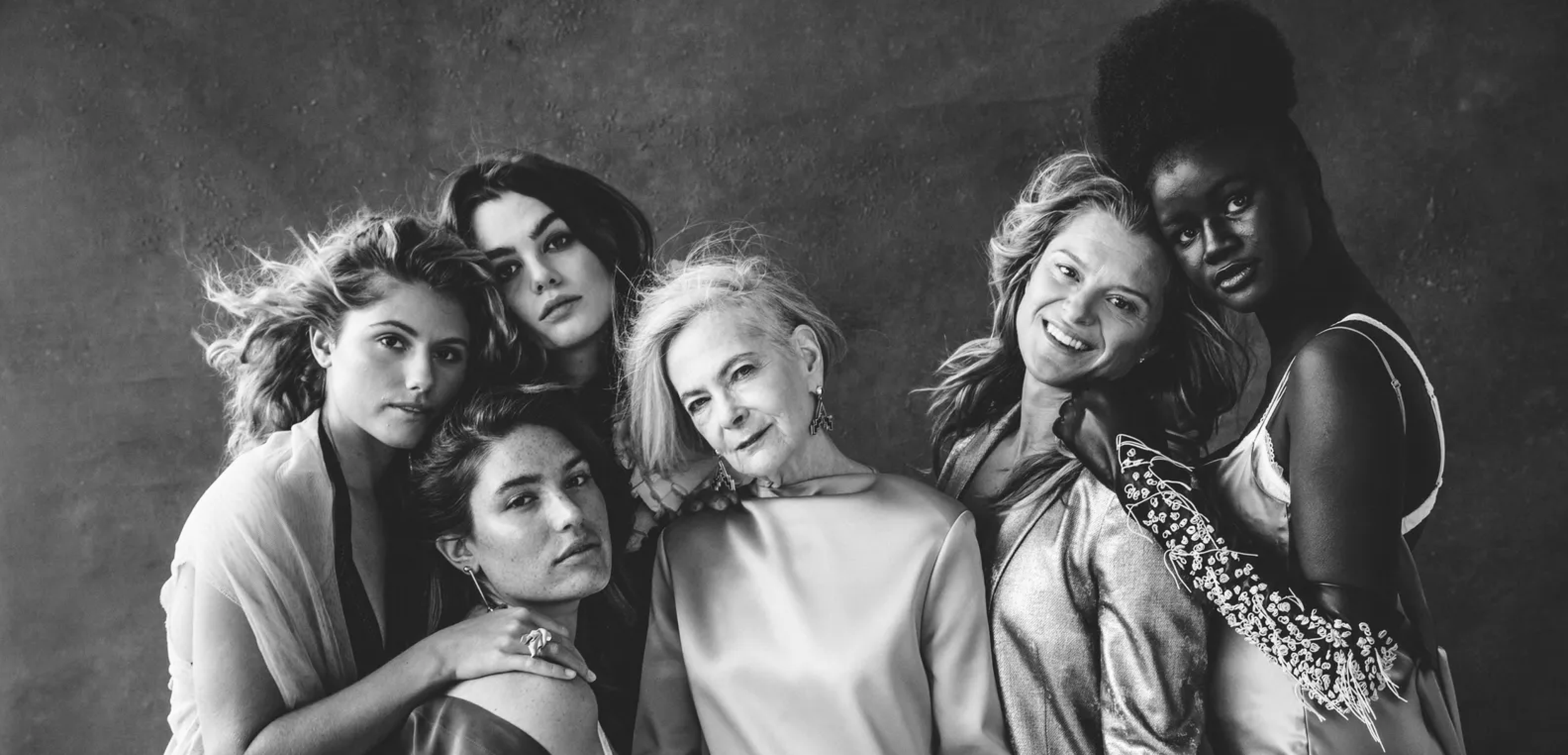 Black and white photo of seven diverse women posing together, some touching each other, in a studio setting.