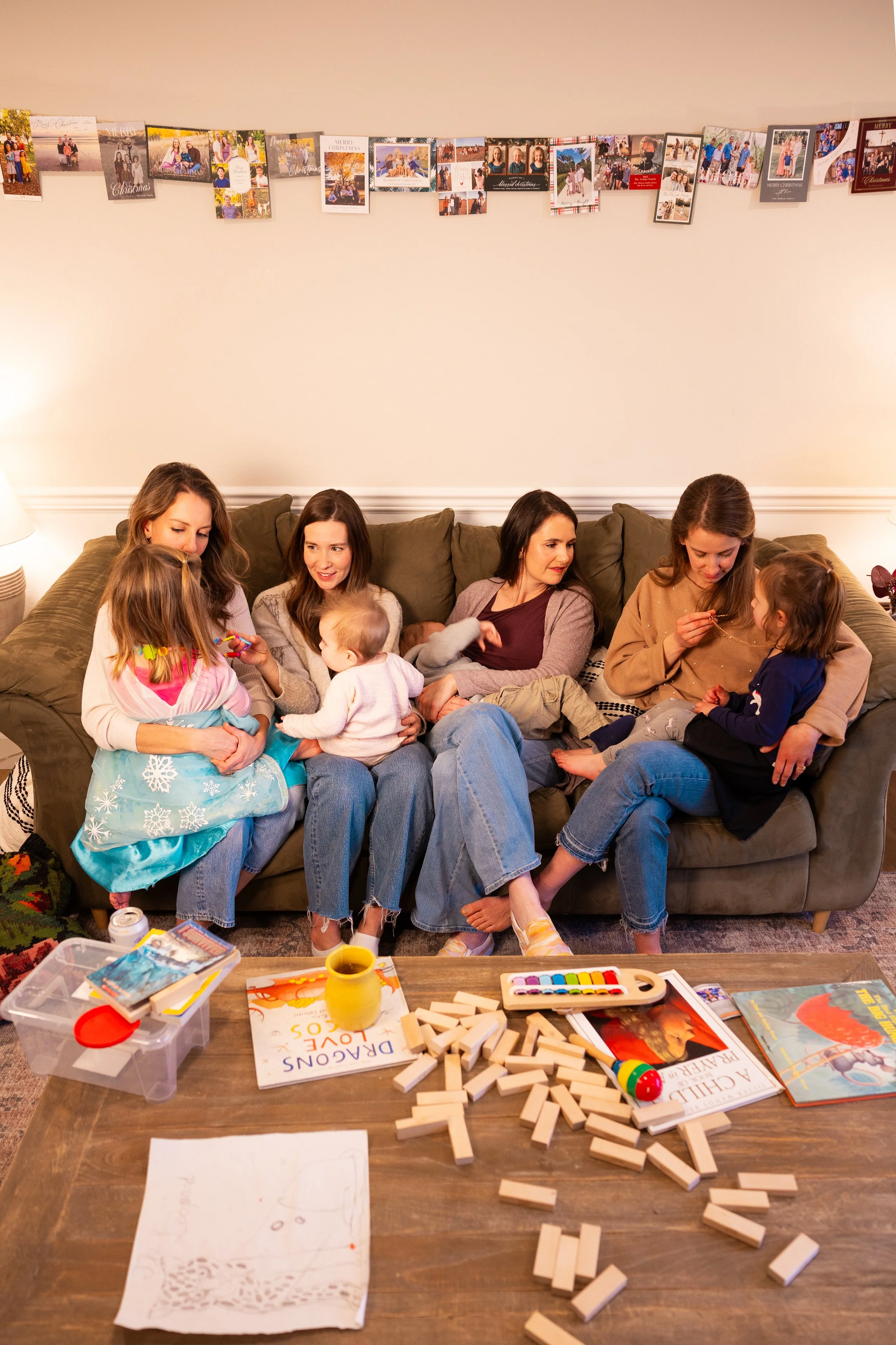 A group of women and children sitting on a couch, engaging in conversation and playing. In front of them, a table holds various board games, books, and craft supplies, including a set of dominoes, a watercolor paint set, and a drawing with the words "DRAGONS LOVE YOU" on it.