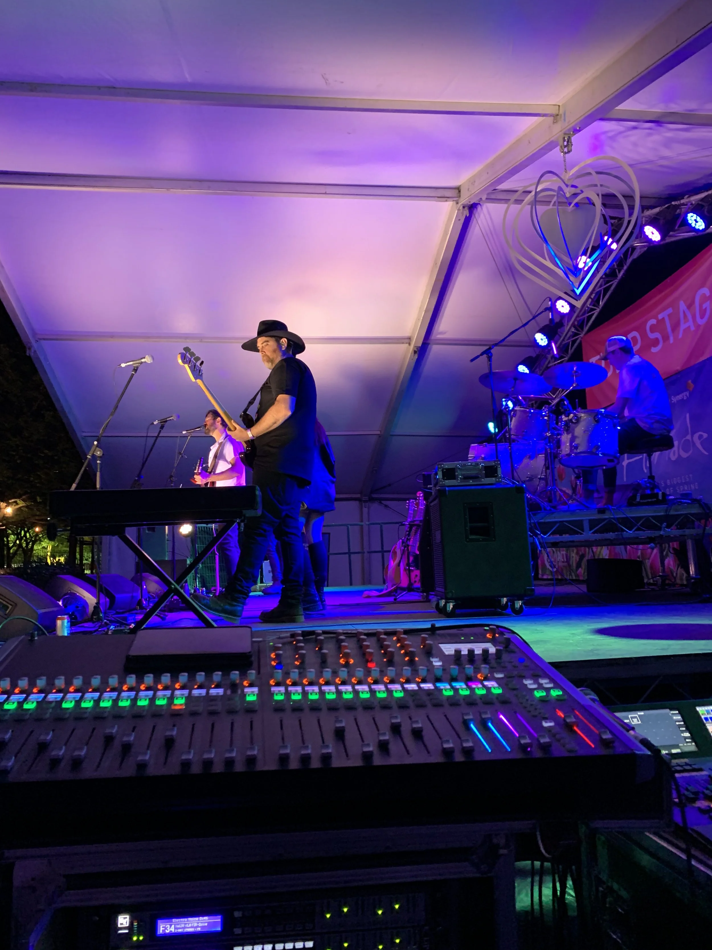 A band performing on an outdoor stage at night, with purple lighting, a heart-shaped decoration hanging above, and a sound mixing console in the foreground.