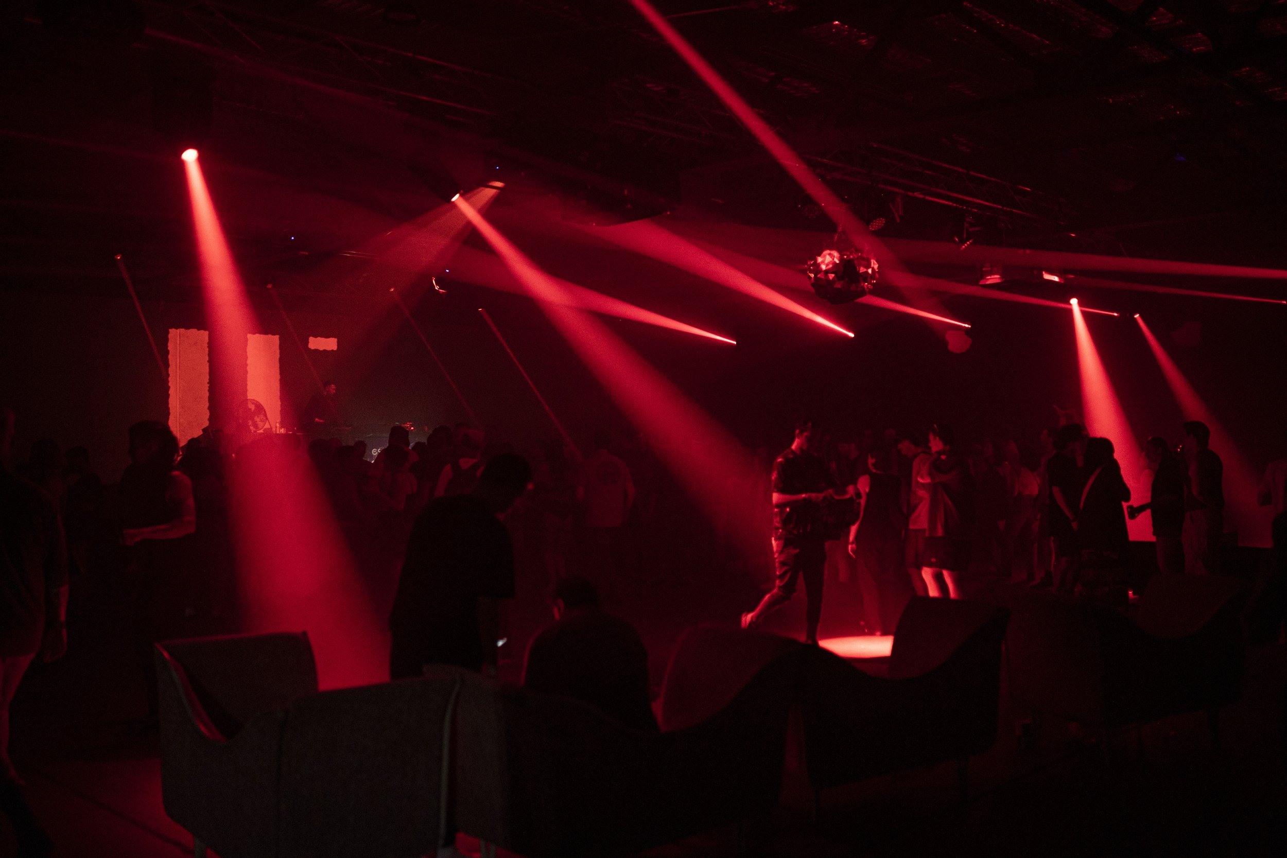 Red stage lighting and spotlights illuminating a dark club or concert venue with a crowd of people.