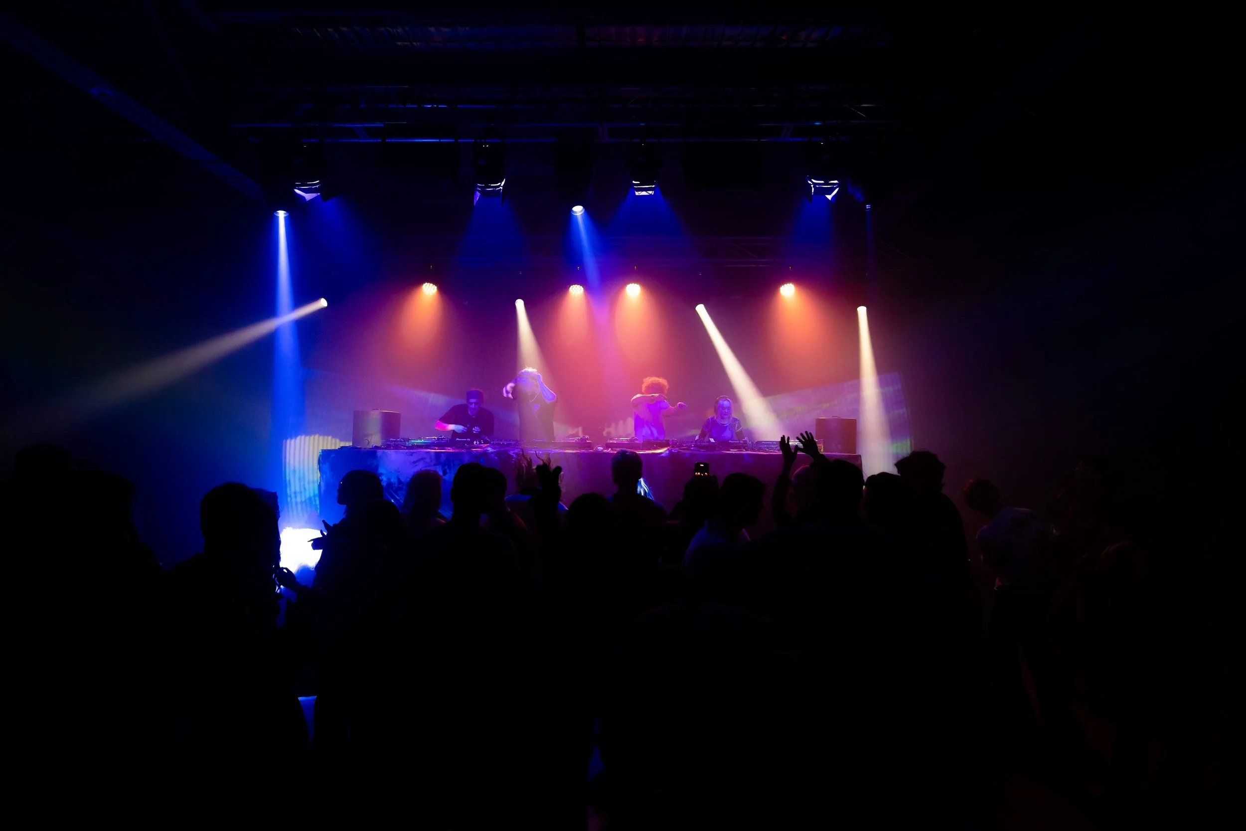 A nightclub with a DJ performing on stage, colorful lighting effects including blue, purple, orange, and yellow spotlights, and silhouettes of the audience with some raising their hands.