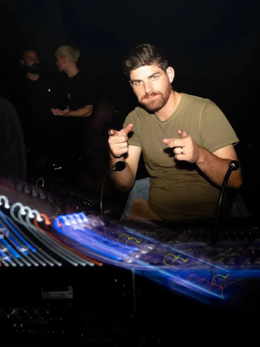 A young man with brown hair and beard pointing at the camera while standing behind a lighting console at a nightclub.