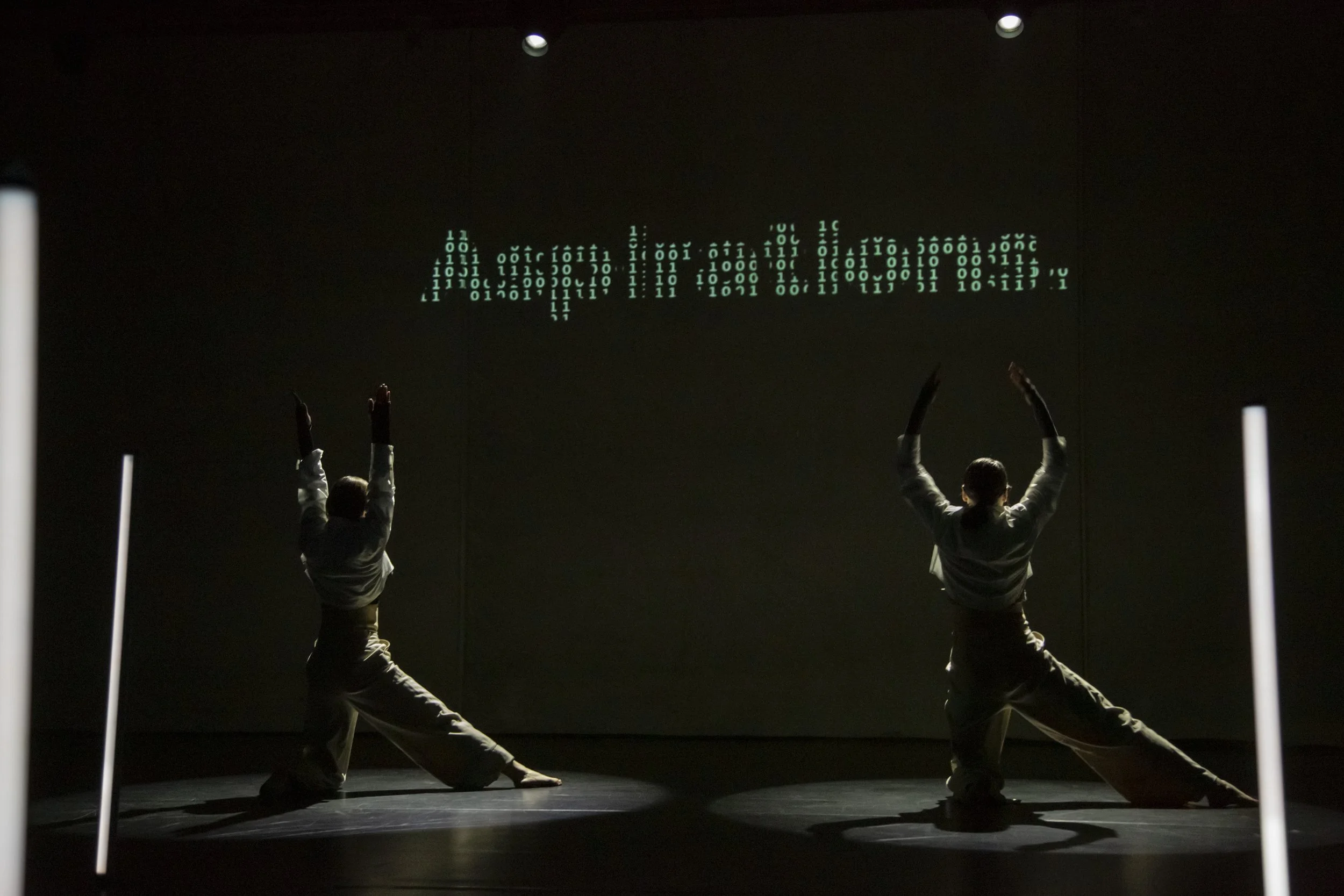 Two performers in black and white outfits dance on stage with their arms raised, silhouetted against a dark background with a line of glowing green binary code forming an abstract shape in the distance.
