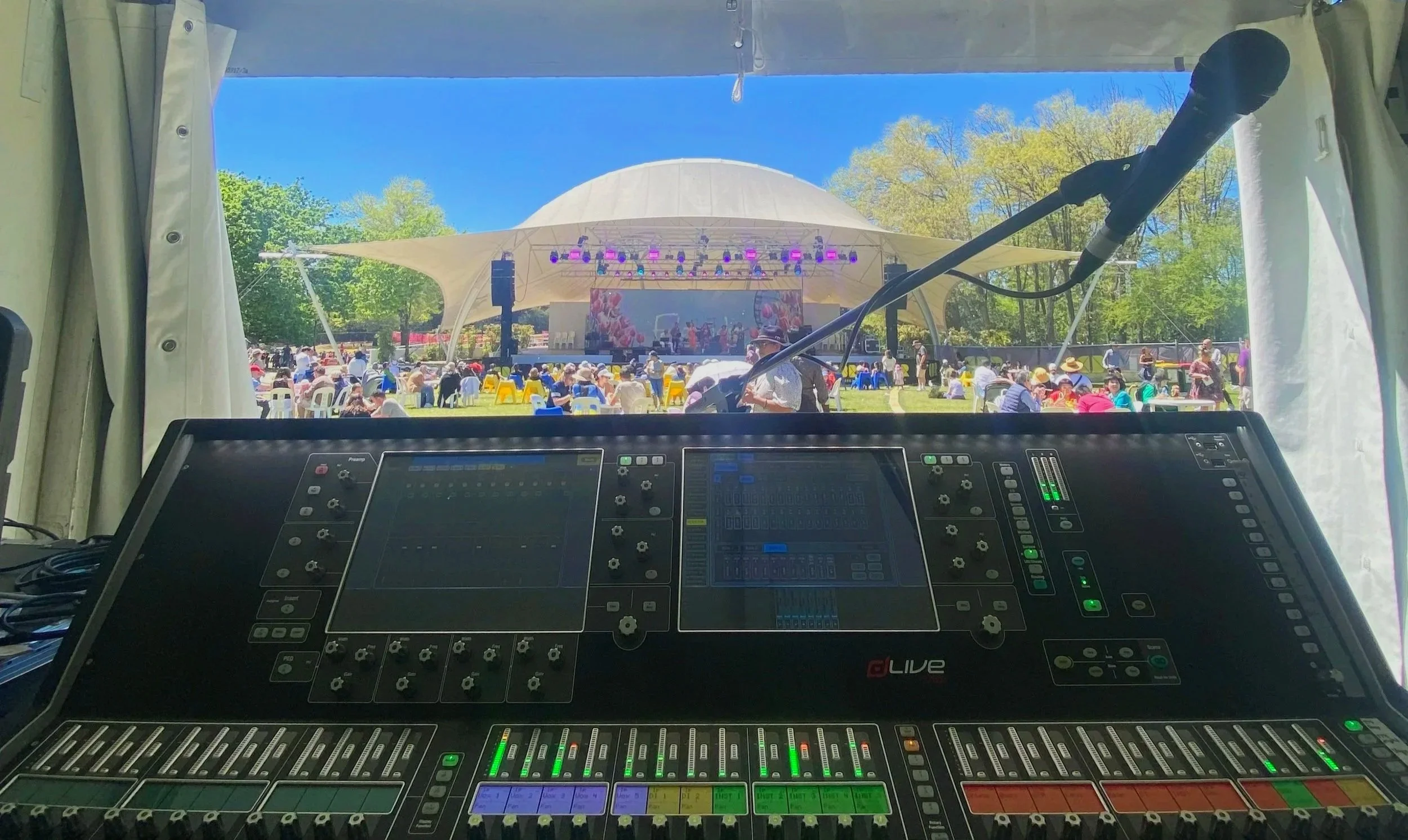 view from inside a tent or sound booth overlooking an outdoor concert stage with people sitting and enjoying the event on a bright, sunny day with clear blue skies and green trees.