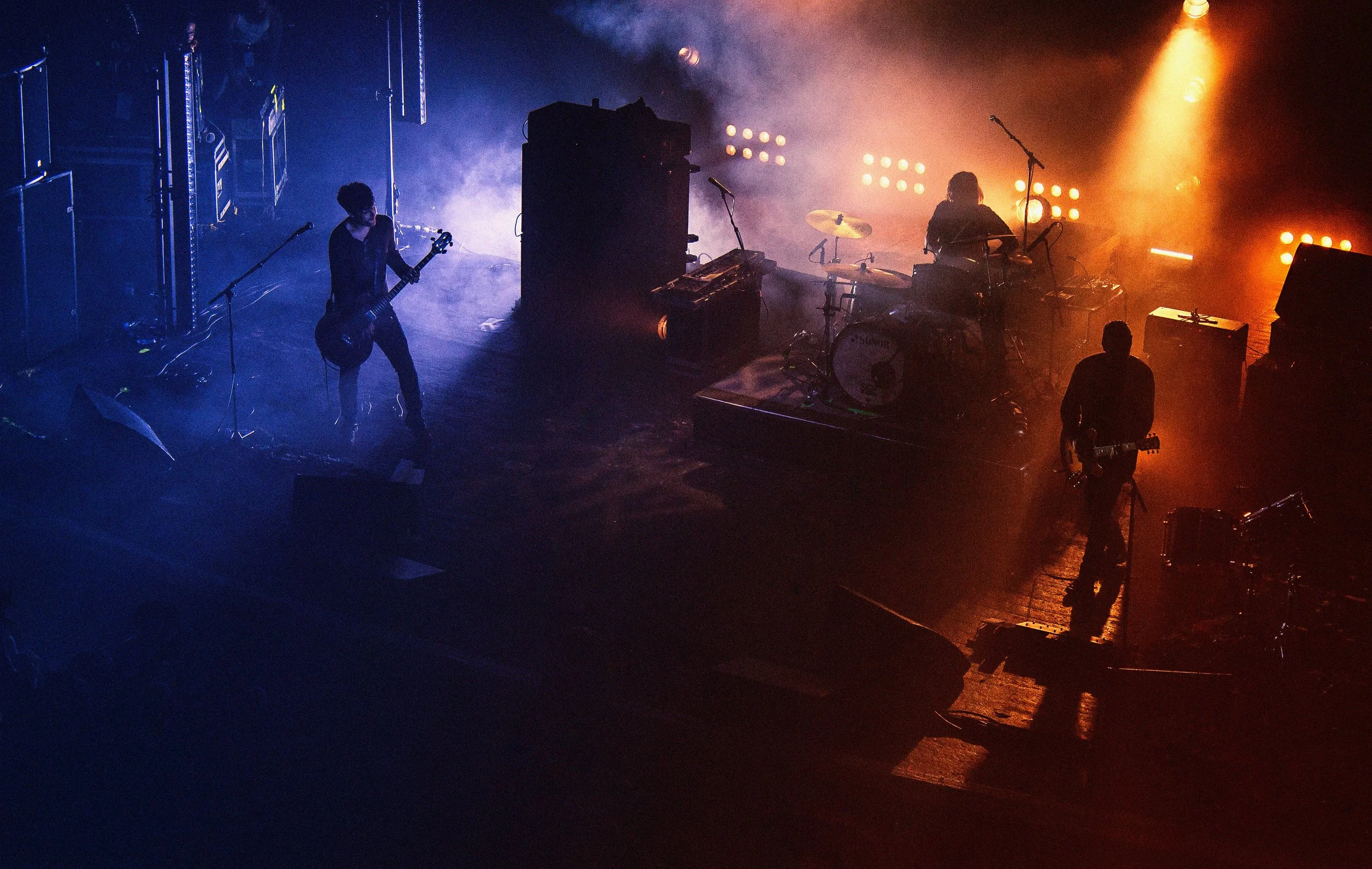 A concert stage with blue and orange lighting, featuring musicians playing guitars and drums, with fog and stage equipment.