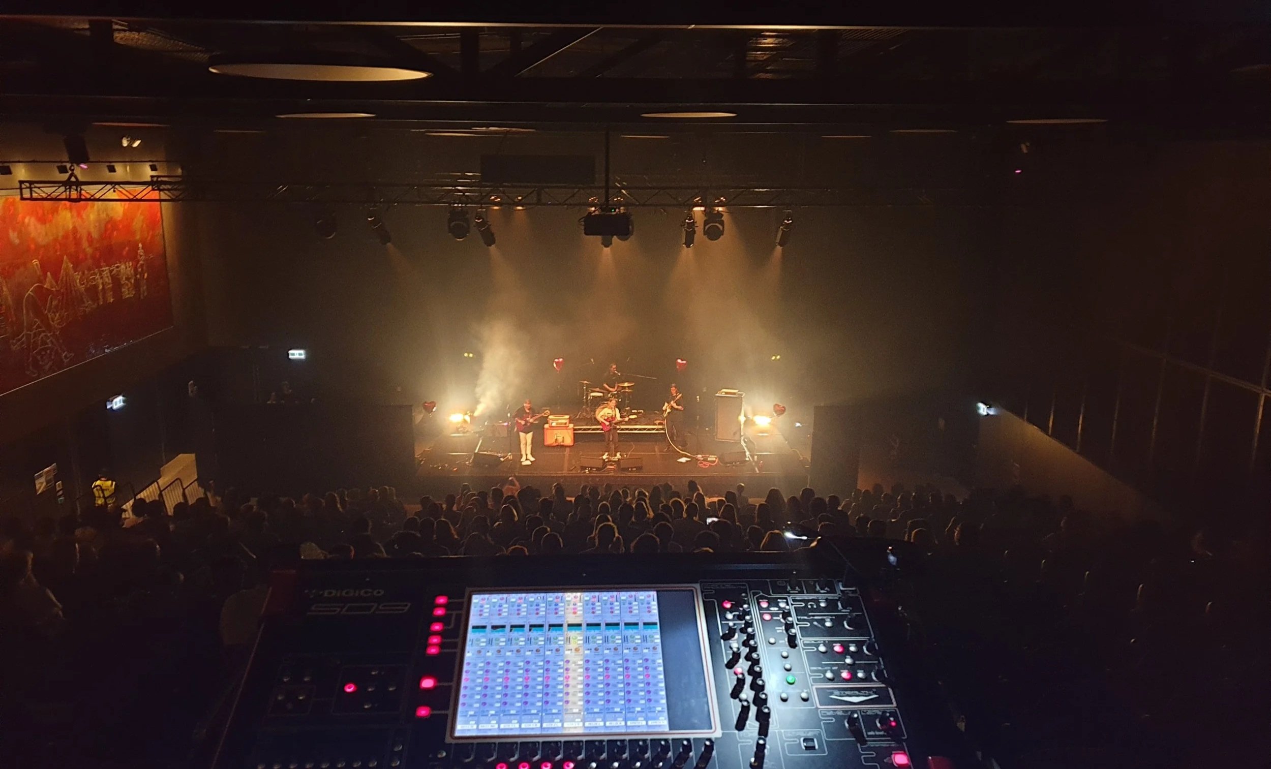 Concert stage with musicians performing, bright stage lights, audience watching, viewed from mixing console in the front.