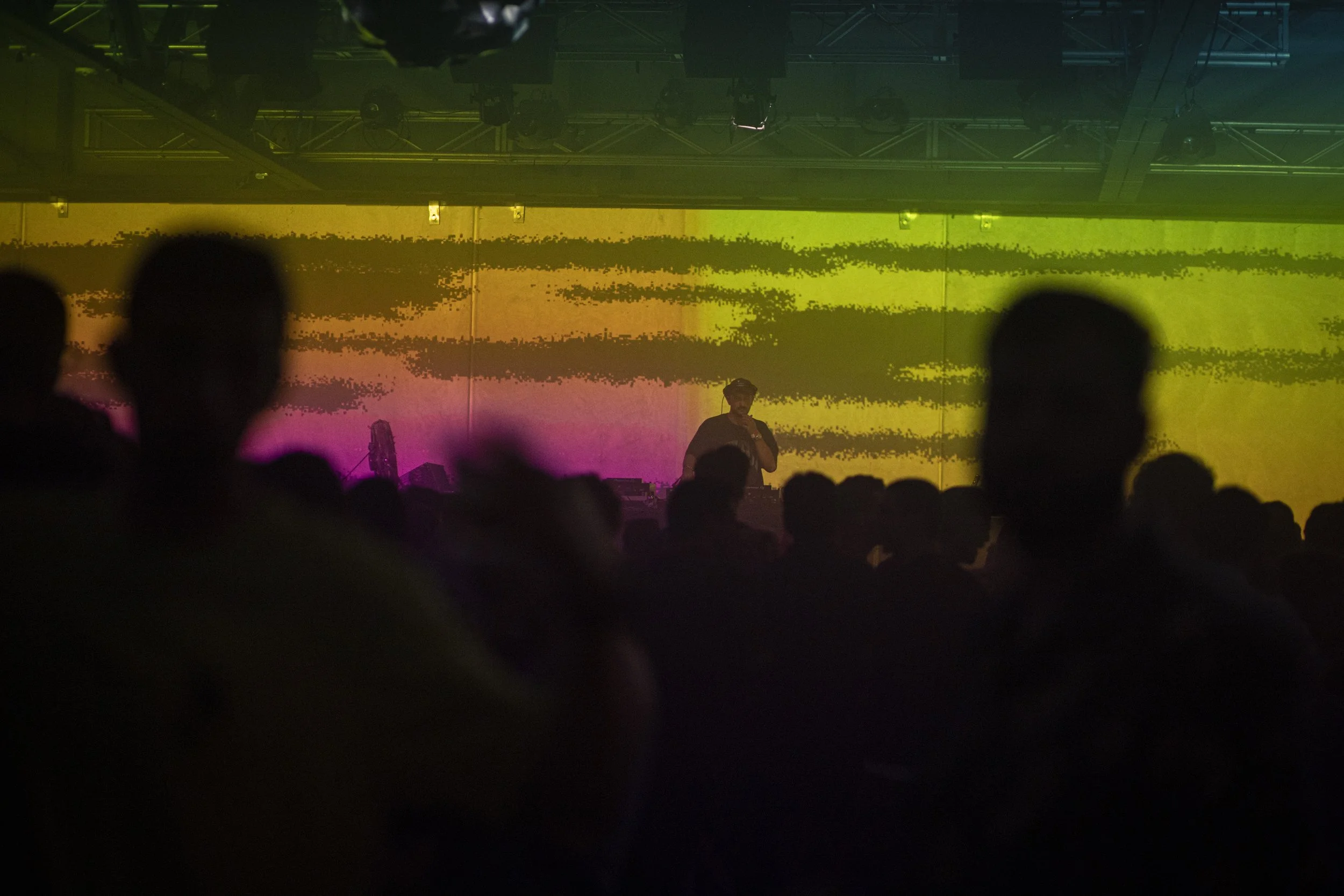 A performer on stage with a rainbow-colored background while an audience watches, with silhouettes visible in the foreground.
