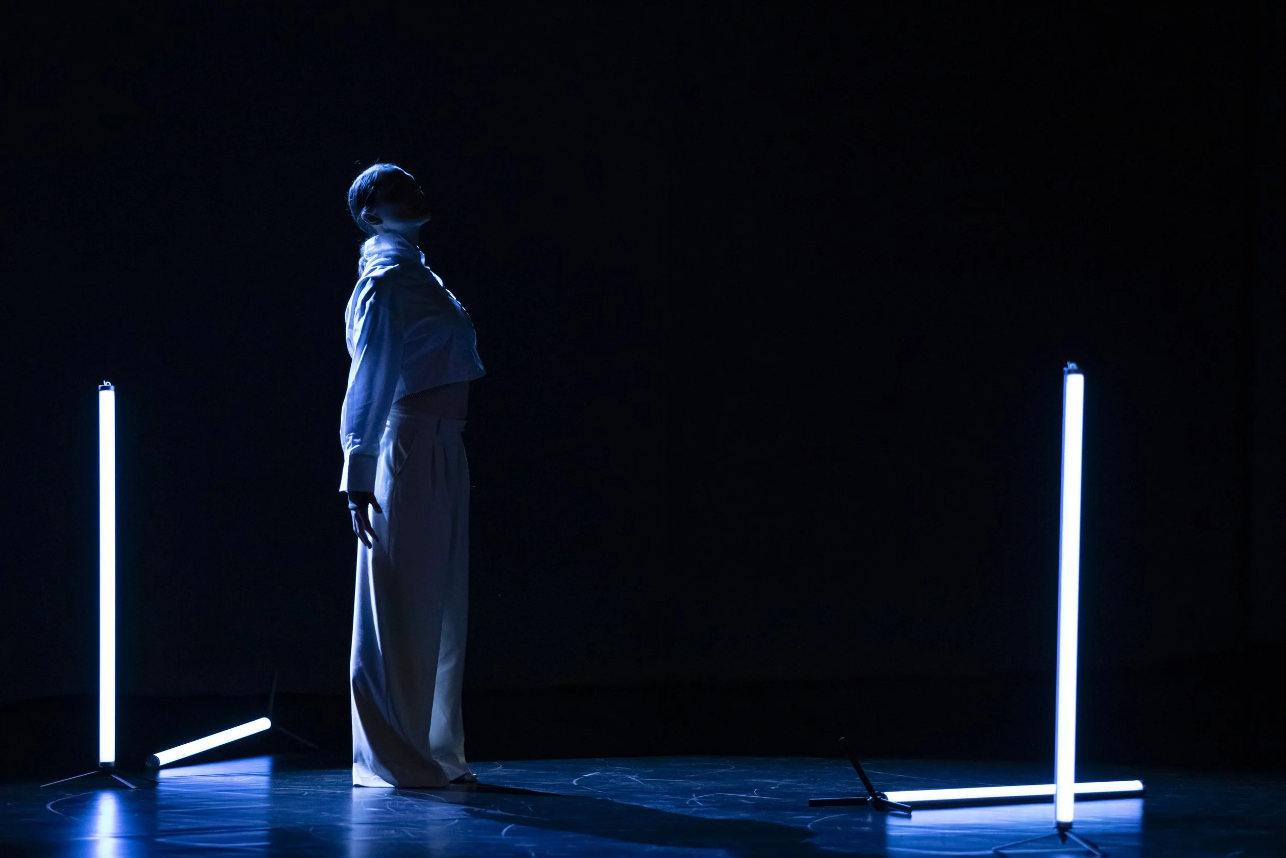 Person standing in a dark space illuminated by blue neon lights on the floor and around the person, creating a minimalistic, futuristic scene.