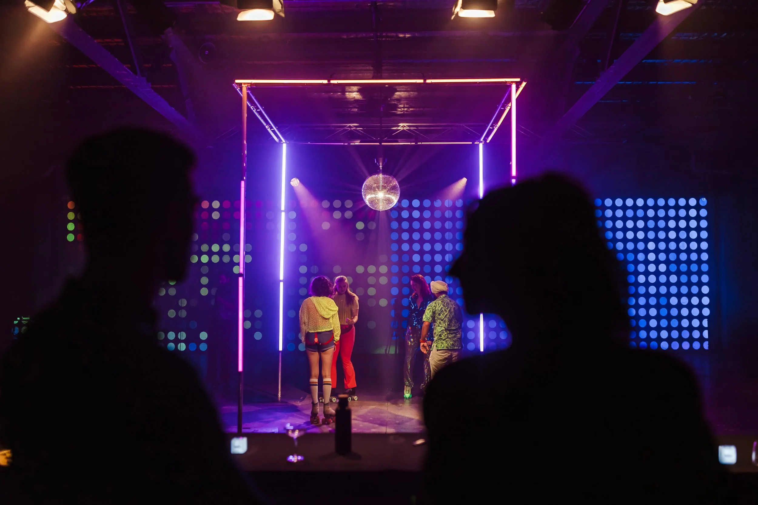 A group of young people dancing and socializing under colorful neon and LED lights on a stage with a disco ball hanging from the ceiling.
