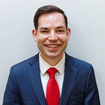 Portrait of a smiling man in a dark blue suit, white shirt, and red tie, standing against a plain light background.