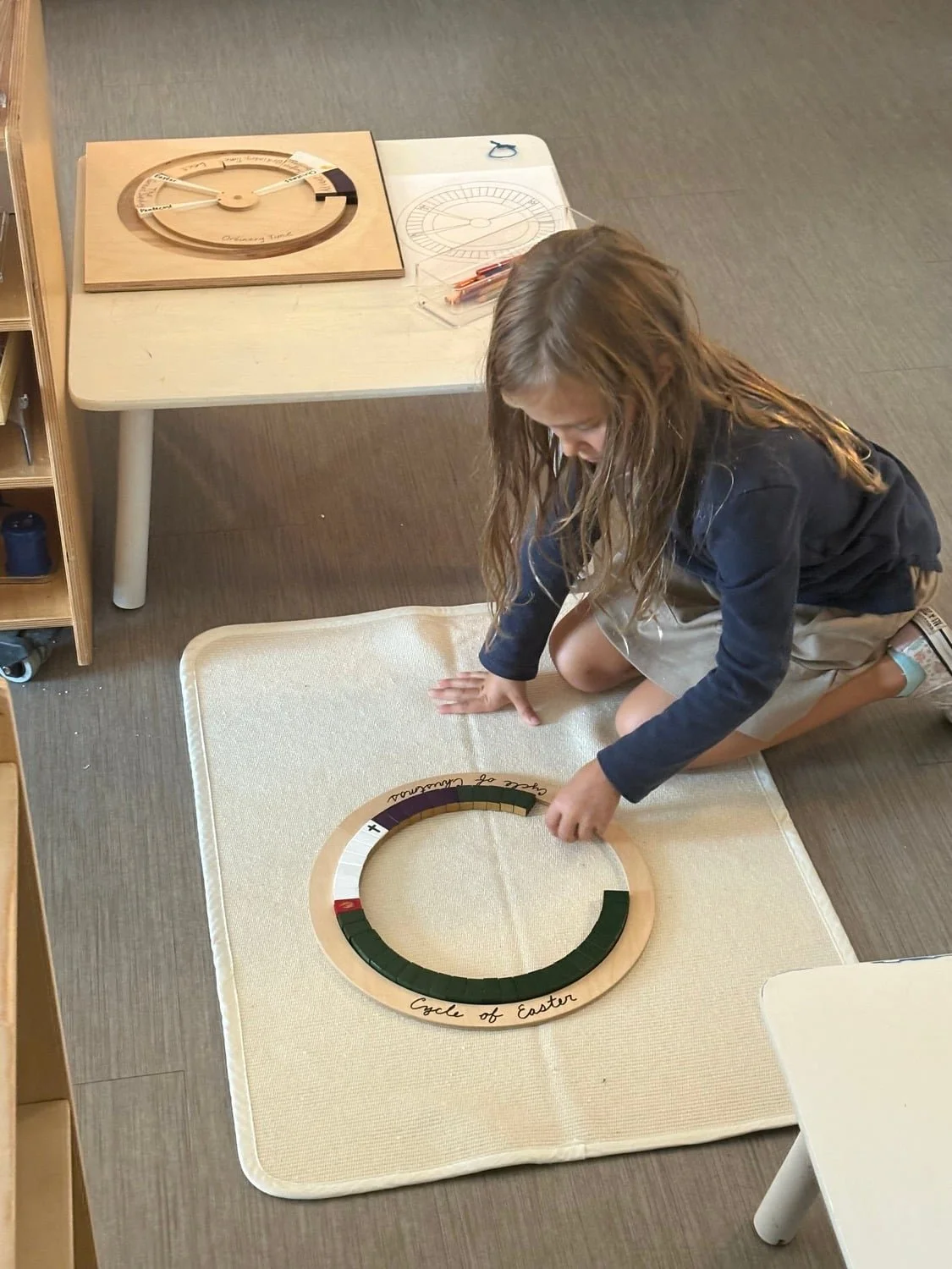 A girl kneeling on a beige mat, placing a colored piece on a circular Montessori material labeled 'Cycle of Easter' and 'Circle of Courage.' A wooden clock and a ruler are on the table behind her.