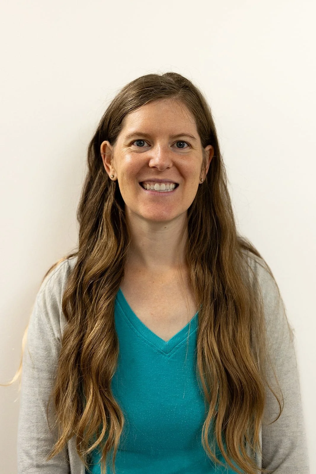 A woman with long wavy brown hair smiling and looking at the camera, wearing a turquoise V-neck shirt and a beige cardigan, standing against a plain white wall.