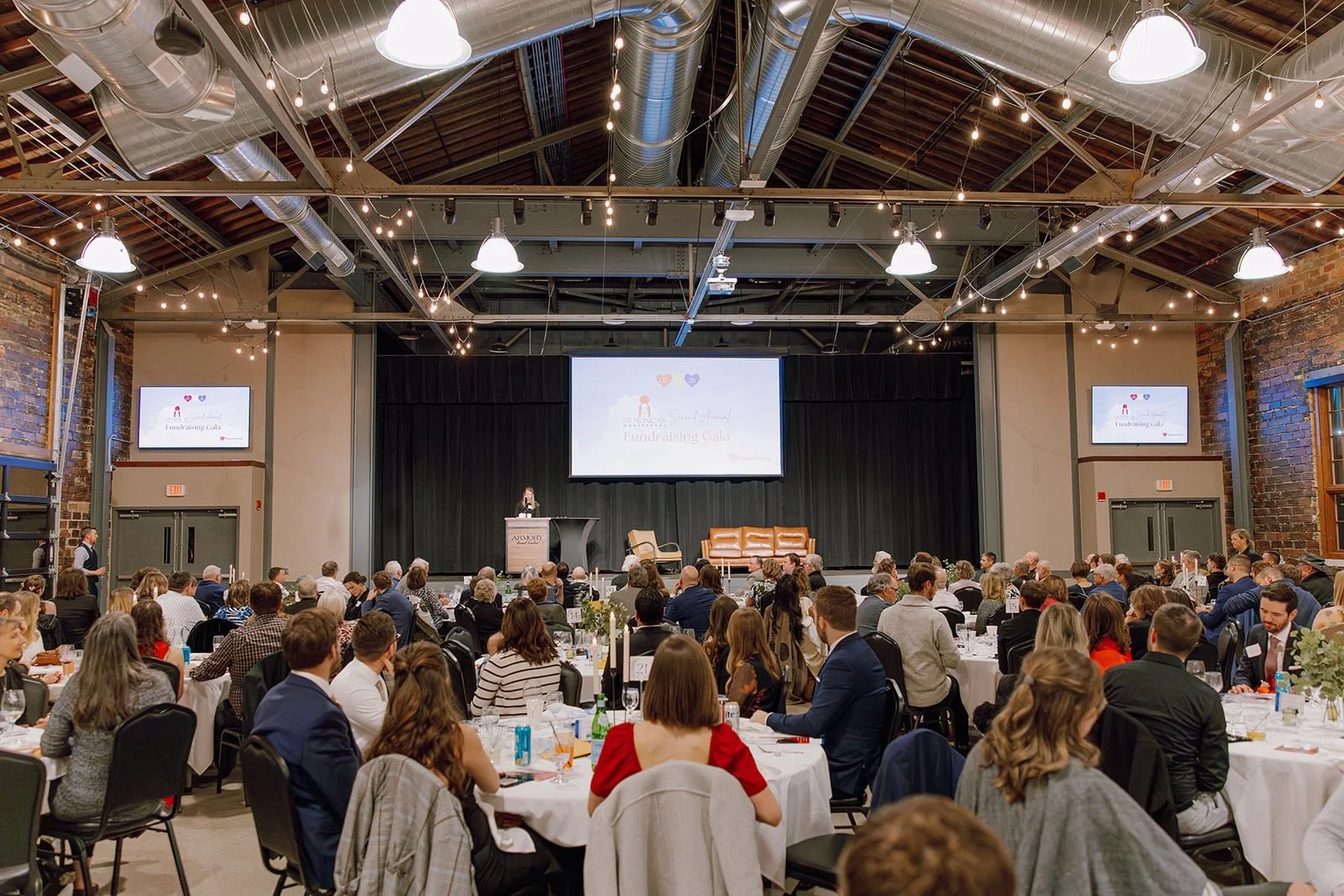 Large indoor fundraising event with round tables filled with seated attendees, a stage at the front, large screens displaying event graphics, and a speaker at a podium. The venue has exposed brick walls, industrial lighting, and a high ceiling with ductwork.