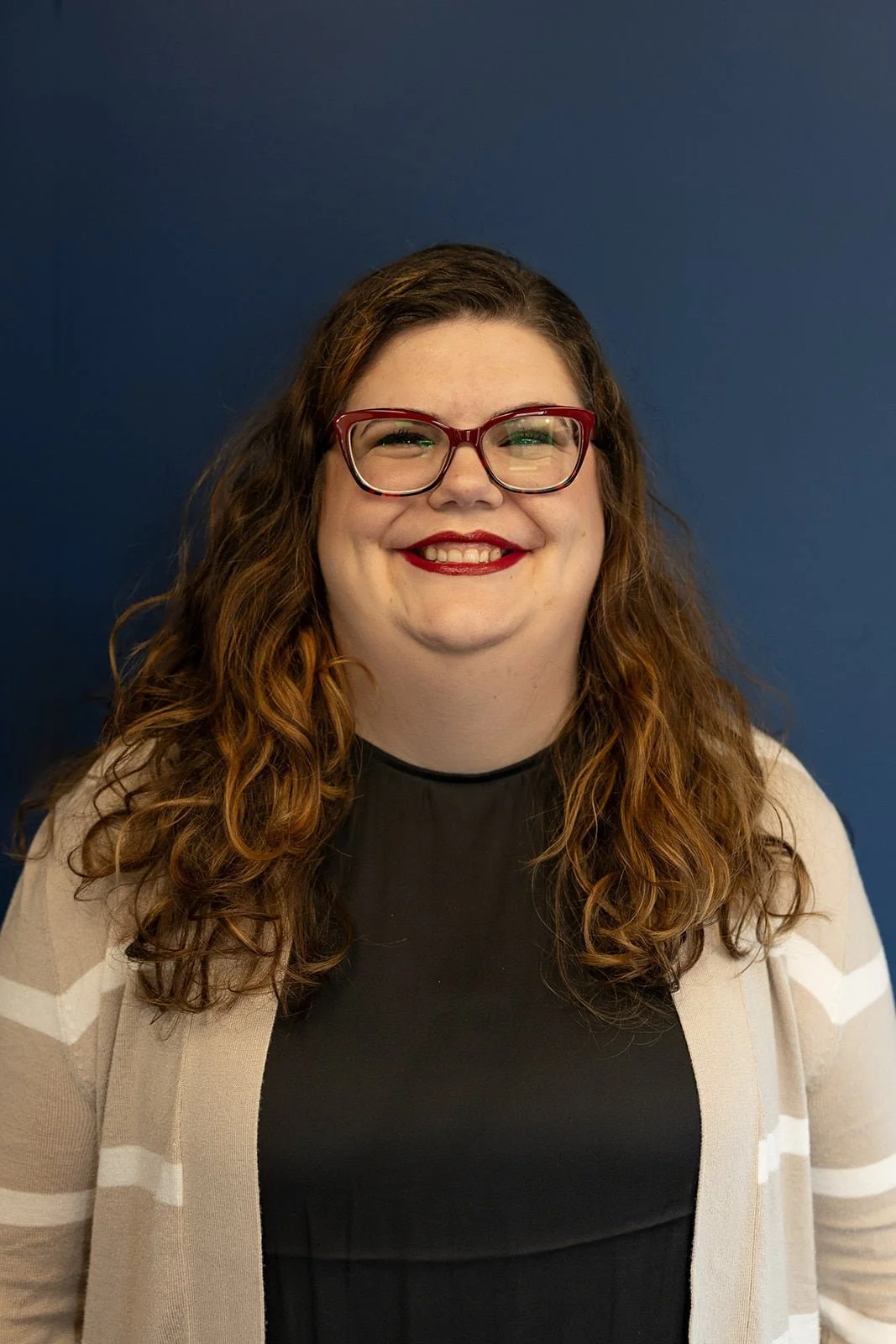 A smiling woman with curly brown hair and red glasses, wearing a black top and a beige cardigan with white stripes, standing against a dark blue wall.