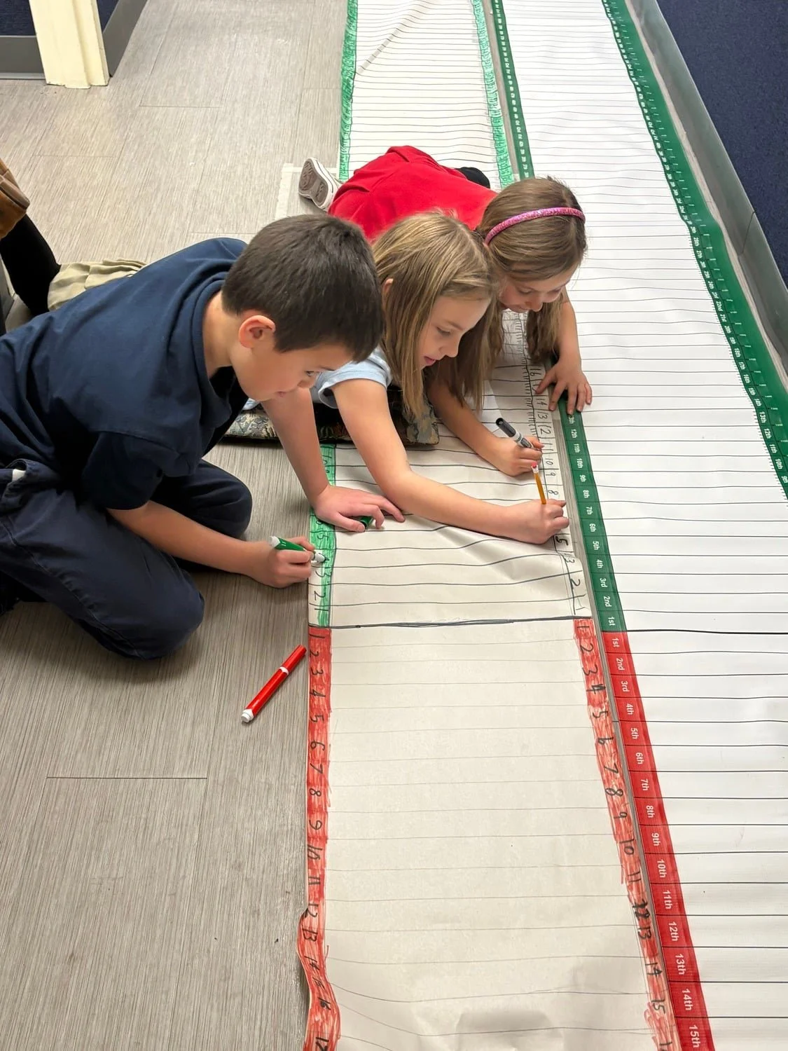 Three children, two girls and one boy, are laying on the floor and drawing on a large educational floor mat that resembles a ruler with red, green, and white sections for measuring height.