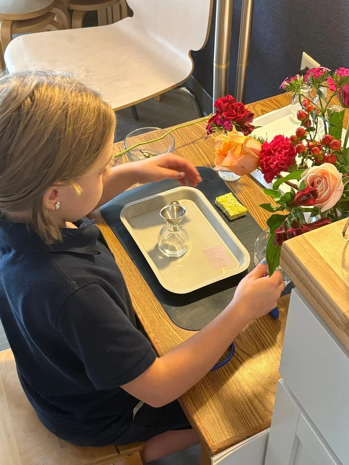 A young girl arranging flowers on a wooden table with a bouquet of pink, red, and peach flowers, a small glass container, a tray, a sponge, and other floral tools.