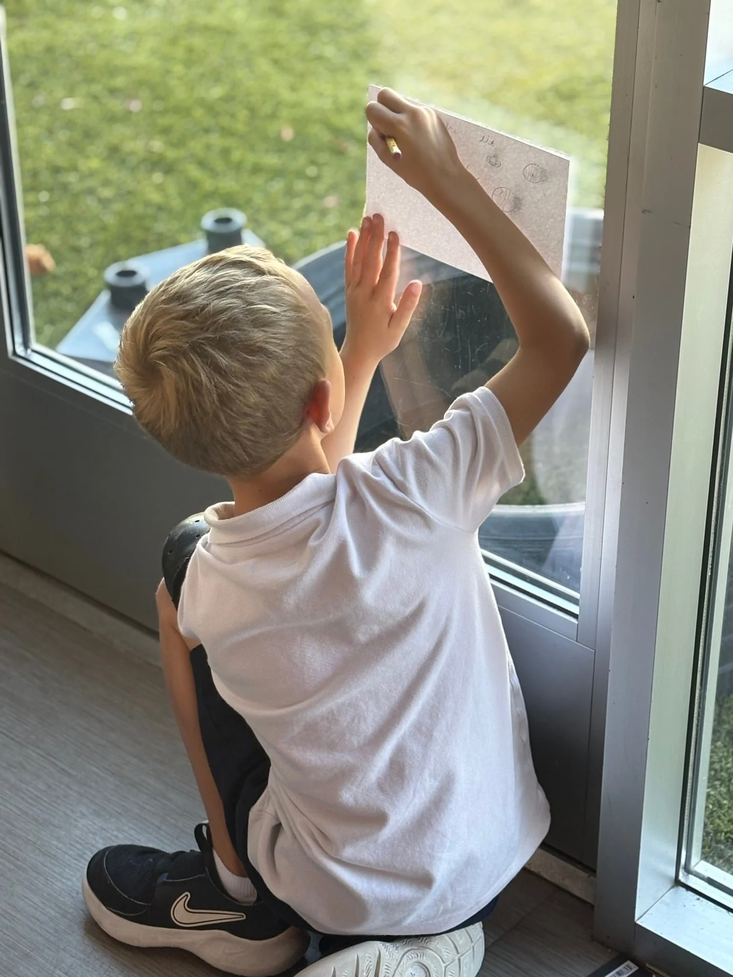 A young boy with blond hair, wearing a white t-shirt, black shorts, and black Nike sneakers, is kneeling on the floor near a window, writing or drawing on a piece of paper taped to the glass.