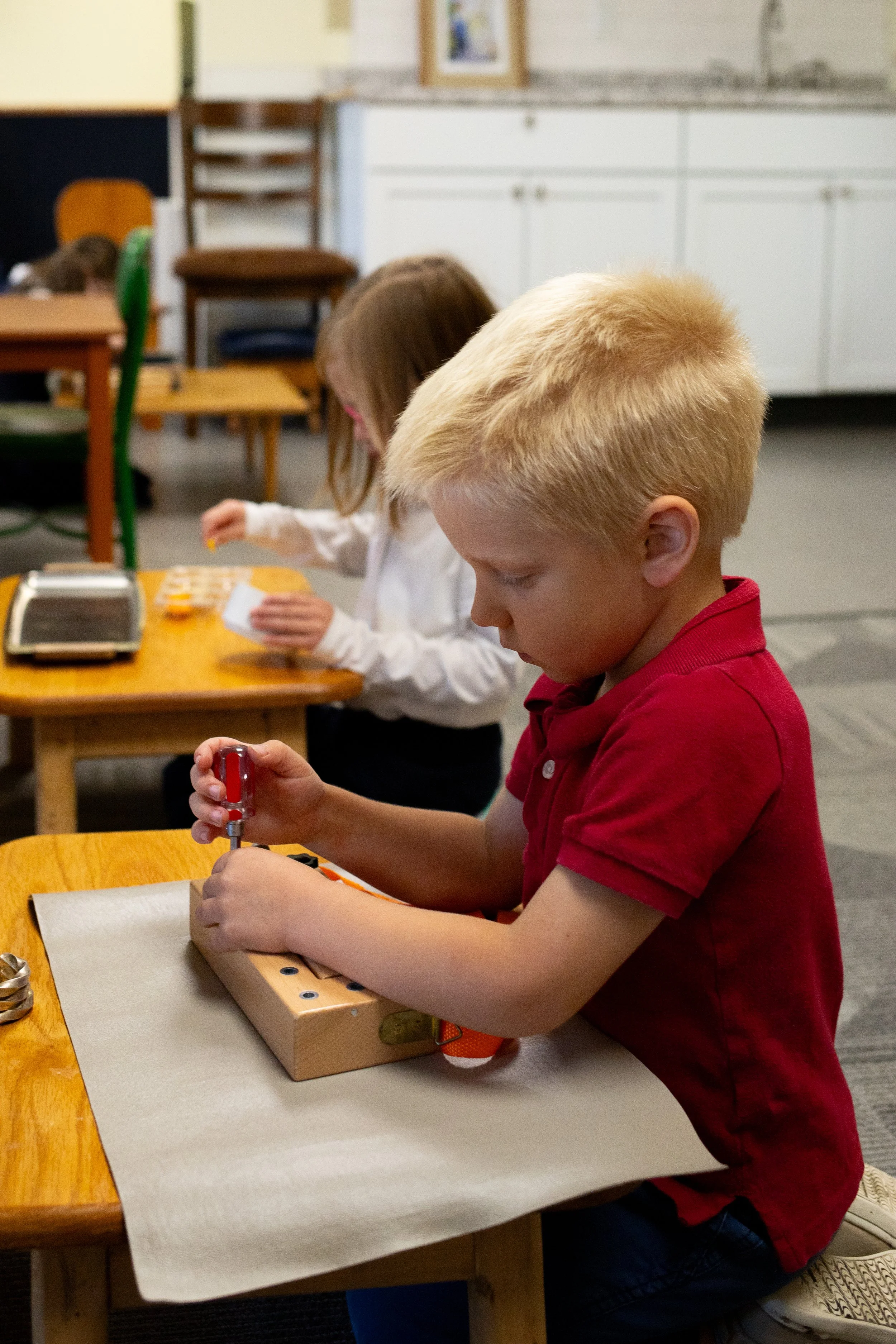 A young blond-haired boy in a red shirt working with a tool on a small wooden device at a table. A girl with brown hair and a white shirt is in the background, also working on a project.