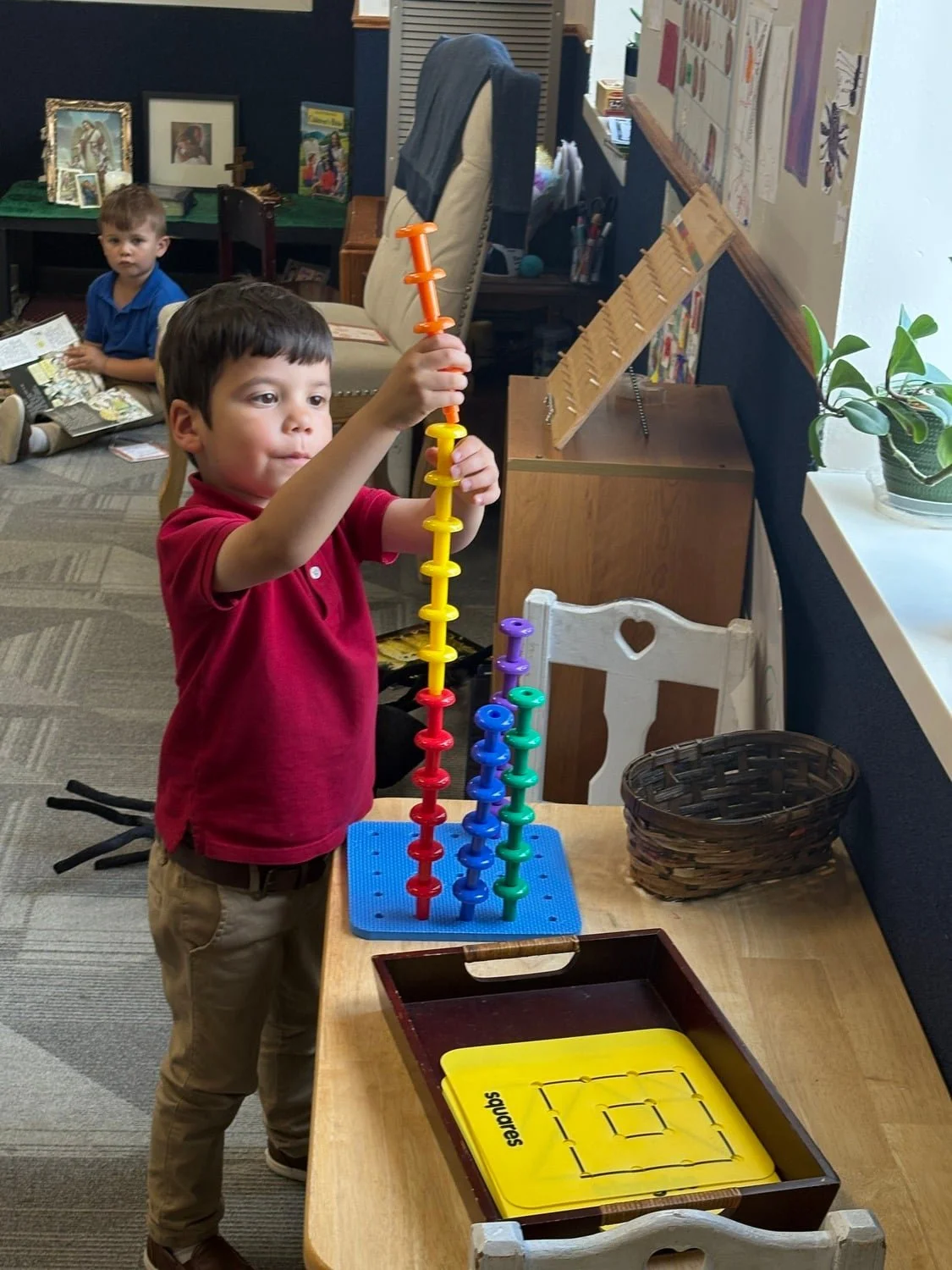 A young boy in a red shirt and khaki pants plays with colorful stacking rings on a table in a classroom, with another boy sitting on the floor in the background reading a book.