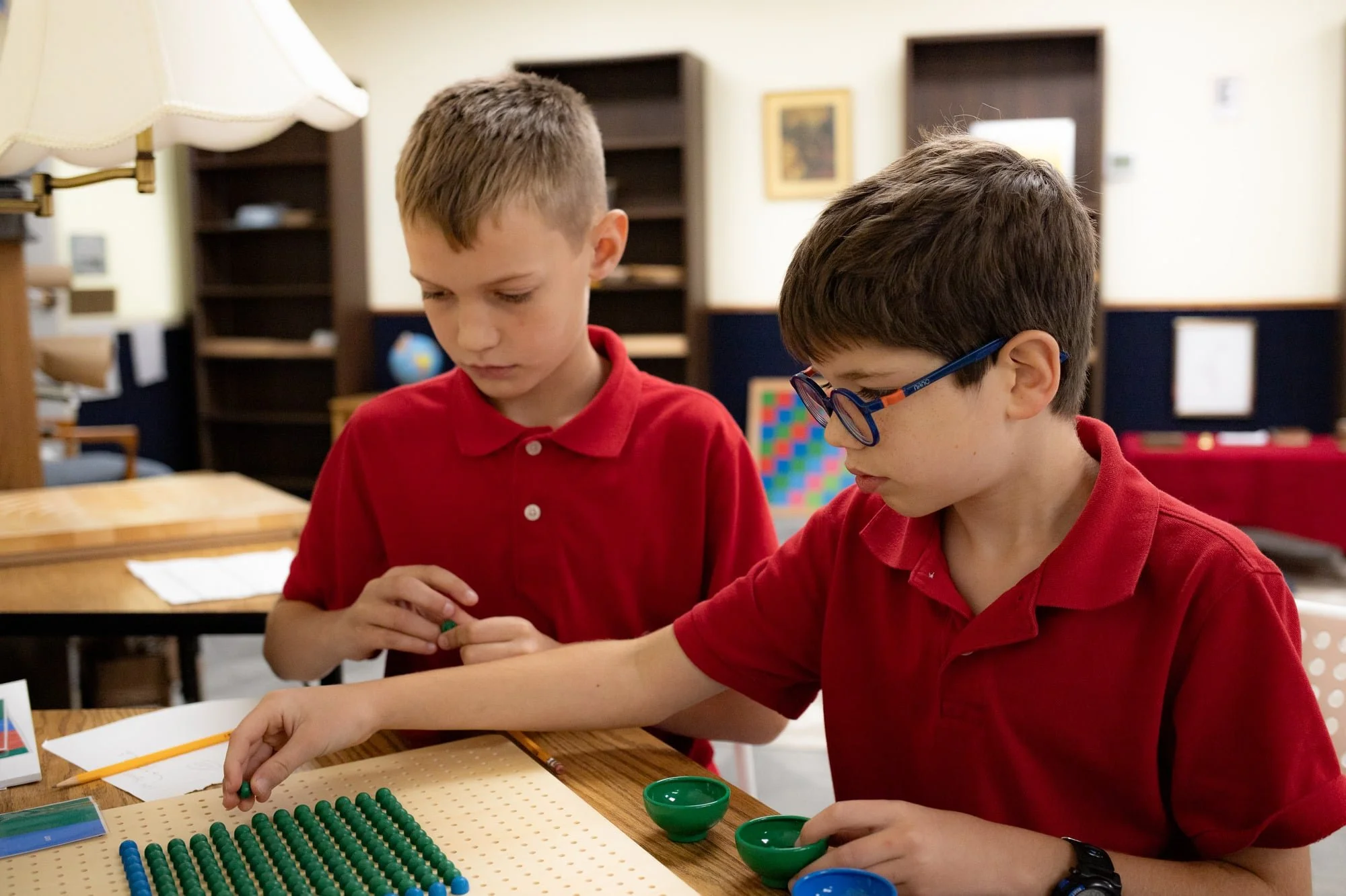 Two young boys wearing red shirts playing checkers game on a table in a classroom.