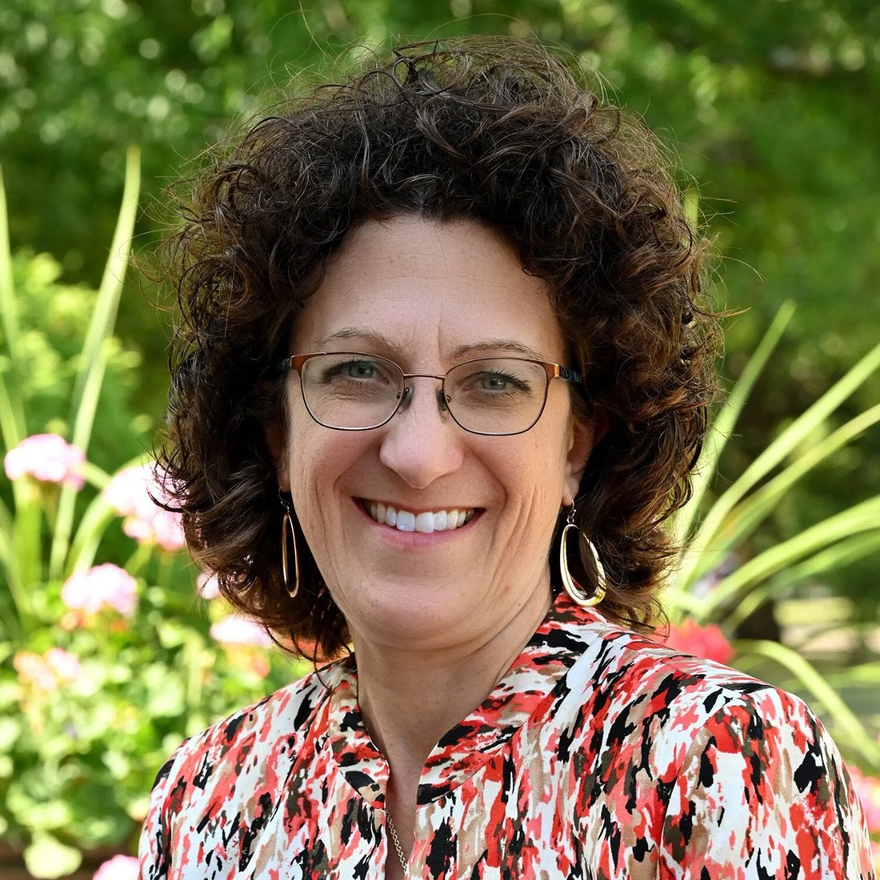 A woman with curly brown hair, glasses, and earrings, smiling outdoors with greenery and flowers in the background.