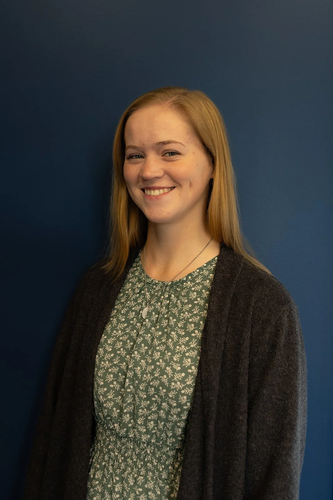 A young woman with red hair smiling, wearing a green floral dress and a black cardigan, standing against a blue wall.