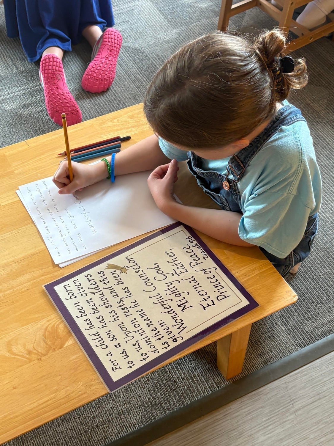 A girl is sitting at a wooden table writing on a sheet of paper with colored pencils. There is a book or poster with handwritten text on the table next to her. Another child is lying on the carpet nearby, wearing pink shoes and dark pants.