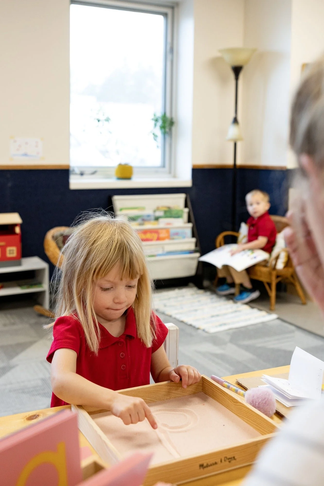 Young girl with blonde hair in a red shirt playing a game on a wooden board at a table in a cozy classroom setting, with a boy sitting in a wooden chair holding a book in the background.