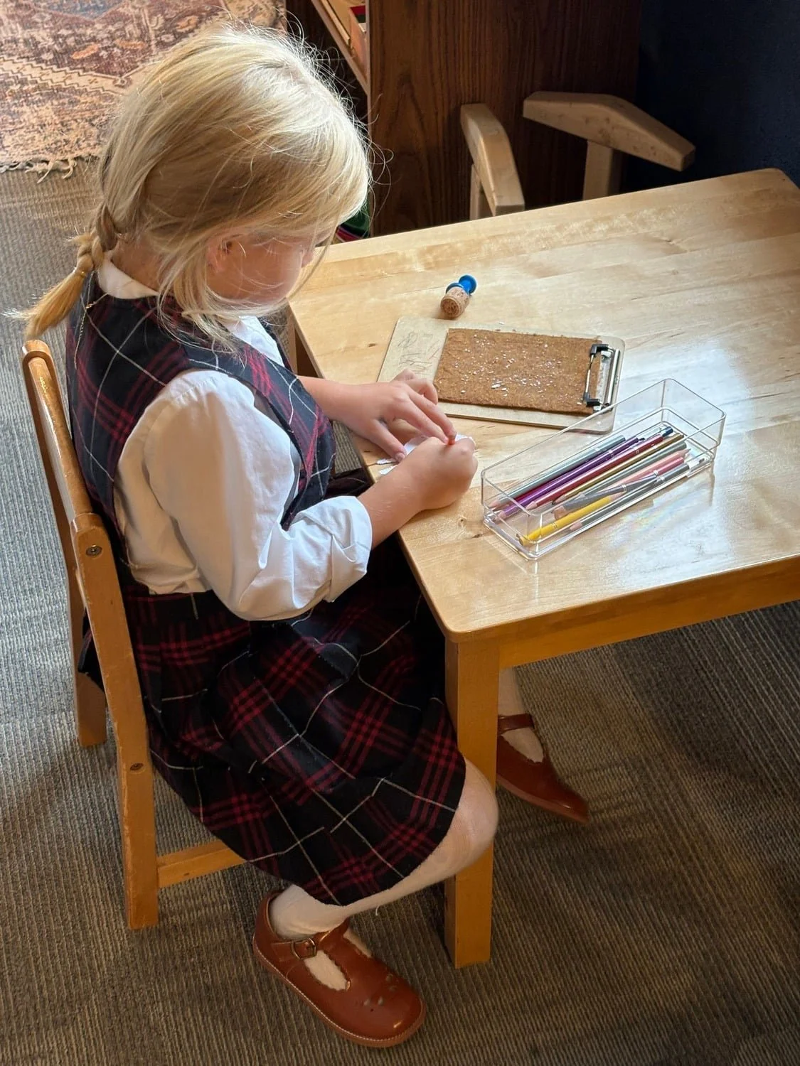 A young girl in a school uniform sits at a wooden table, working with craft materials including colored pencils and a corkboard.