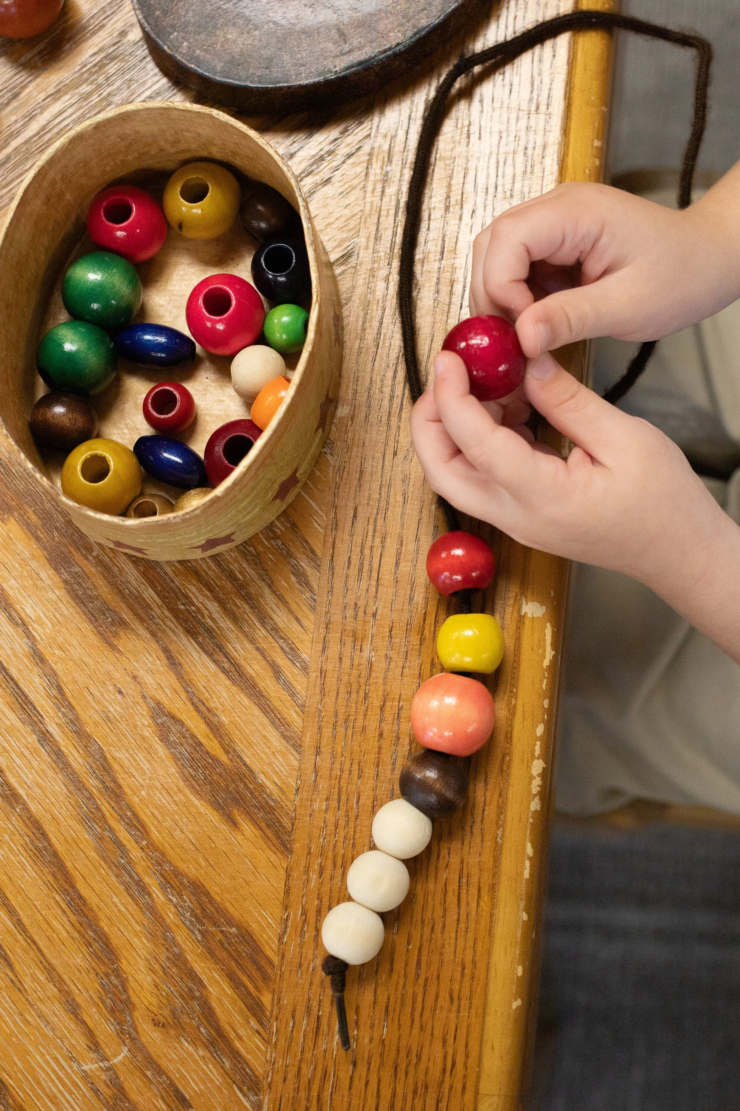A person holds a red wooden bead string on a wooden table, with a cylindrical container filled with colorful beads nearby.