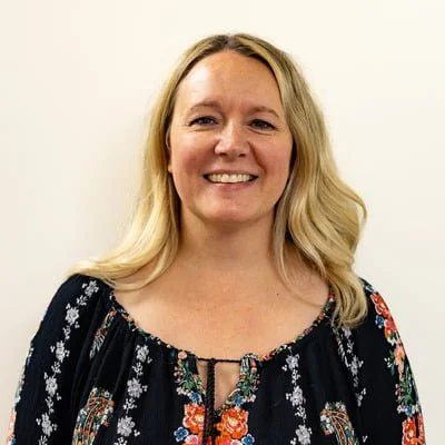 A woman with blonde hair smiling, wearing a dark floral top, standing against a plain light background.