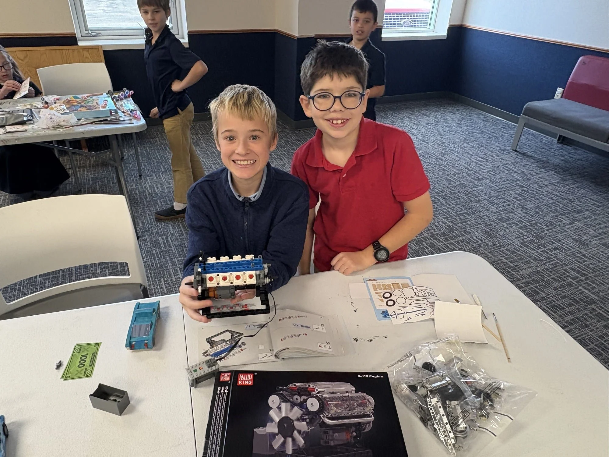 Three boys smiling at the camera with a LEGO robotics project on the table. The boy on the left has blonde hair and is holding a LEGO structure, the boy on the right has dark curly hair and glasses, and the boy in the background has short dark hair. There are LEGO kits, instruction manuals, and other building materials on the table, and a window in the background.