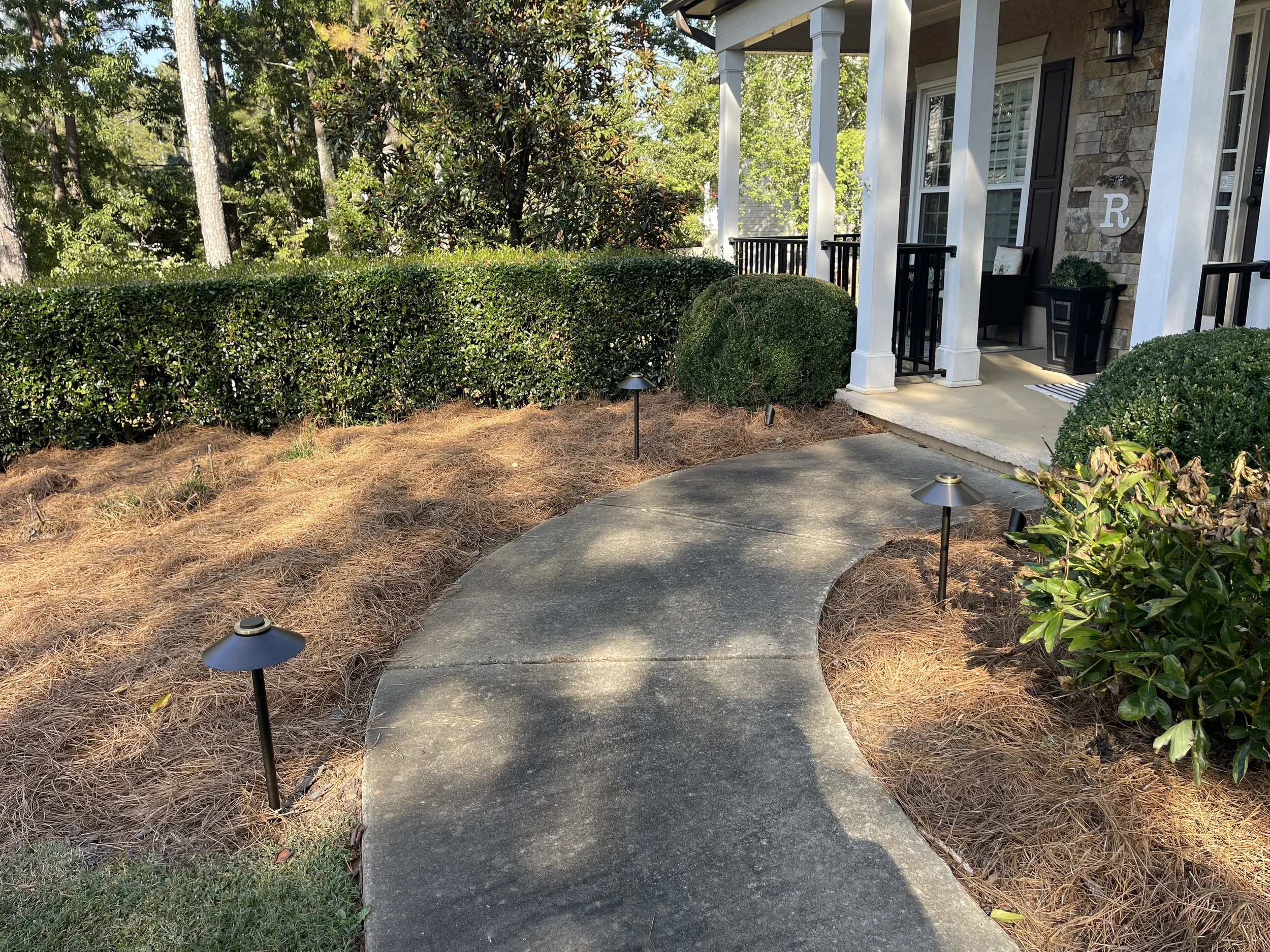 A sidewalk leading up to the front porch of a house, with small landscape lights along the walkway, bushes, trees, and part of the house's front porch and wall visible.