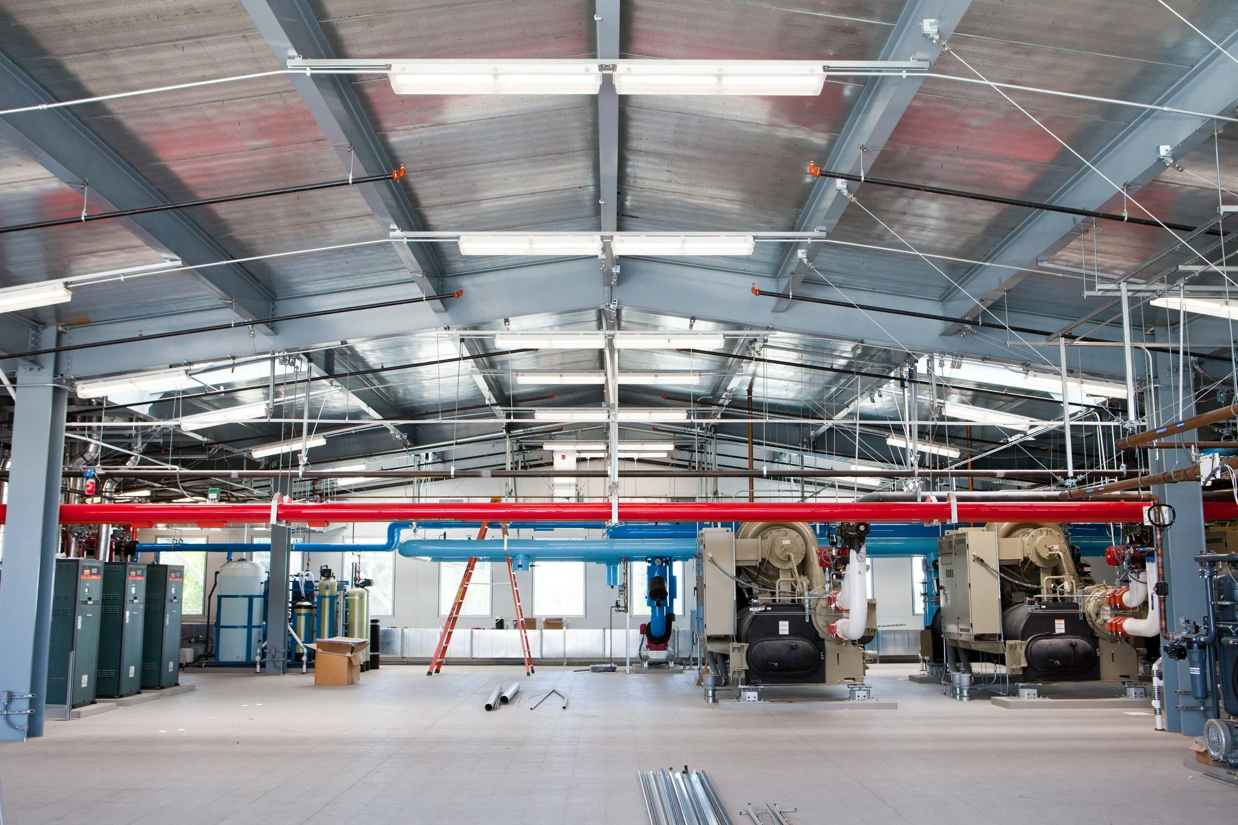 Industrial facility interior with exposed metal roof, piping, machinery, and equipment, including large ventilation and air handling units, with ladders and construction materials in the space.
