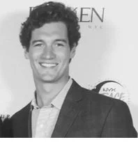 A young man with curly hair smiling, dressed in a suit, at an event or photo session.
