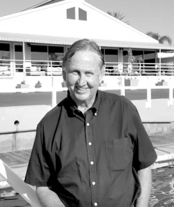 A smiling man standing outdoors in front of a building with a porch, near a body of water.