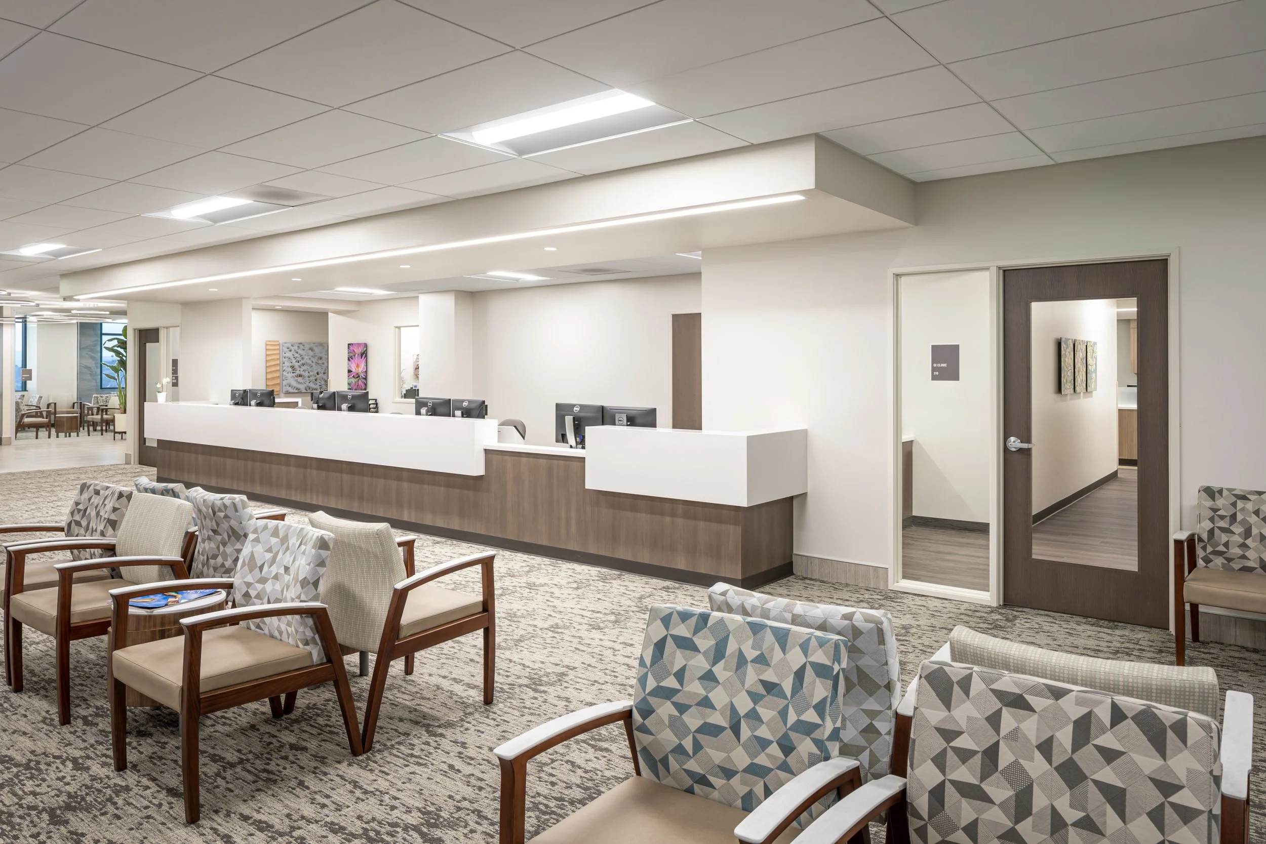 A hospital or clinic reception area with a row of chairs with wooden arms and patterned upholstery. There are paintings on the wall behind the reception desk. The reception desk has computers on it. The ceiling has recessed lighting, and there is a glass door on the right leading to another room. In the background, there is a waiting area with more chairs, tables, and plants near the windows.